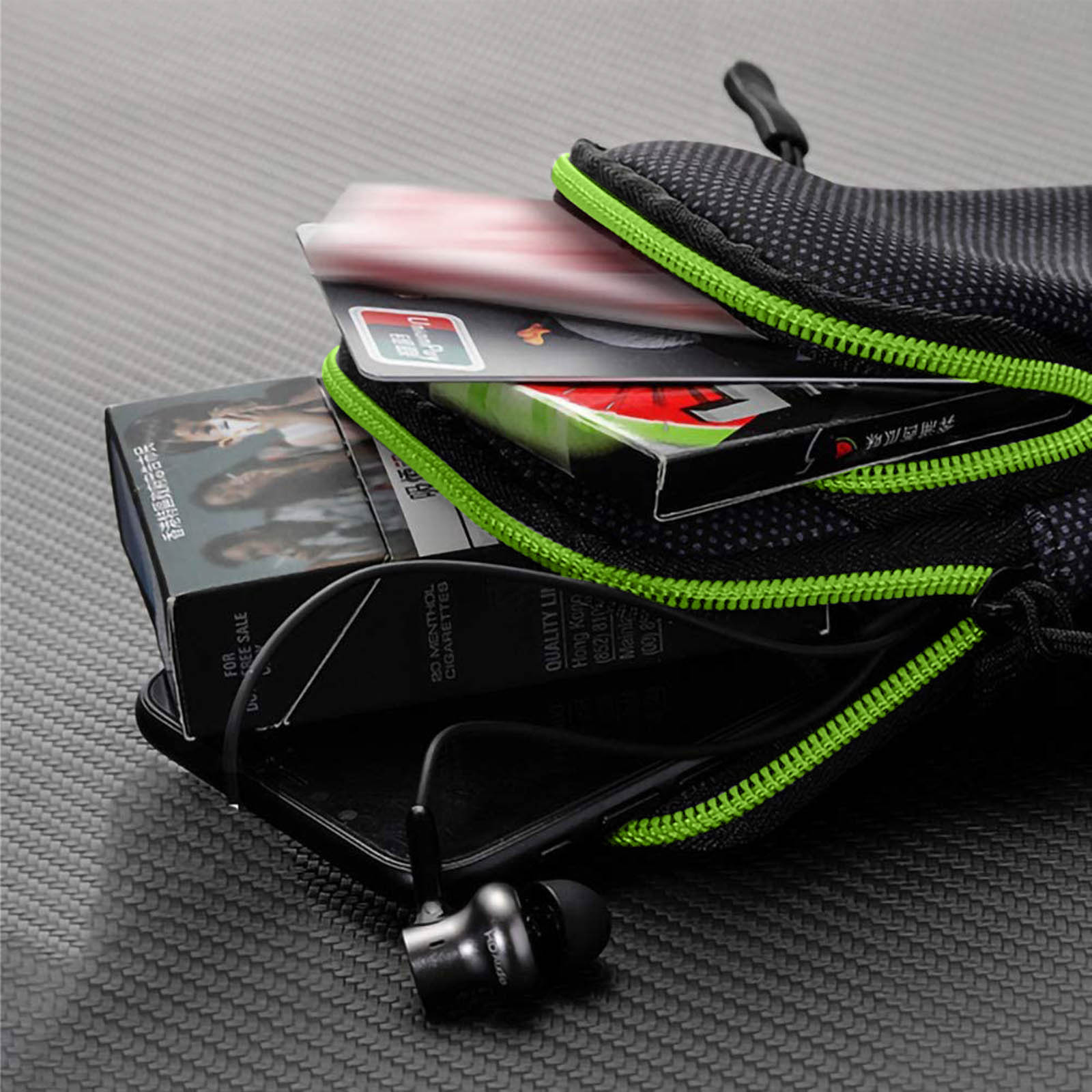 Une pochette noire avec fermeture éclair verte contient des cigarettes, des cartes et des écouteurs. La pochette est sur une surface texturée.