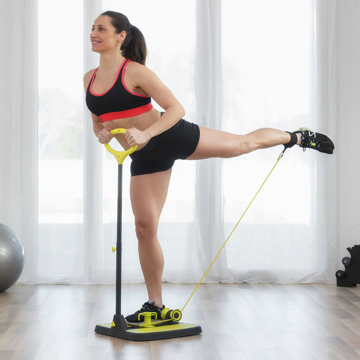 Mujer haciendo ejercicio con una máquina de fitness. Está en un gimnasio, levantando la pierna. La máquina es amarilla y negra. Lleva pantalones cortos y un top negros.