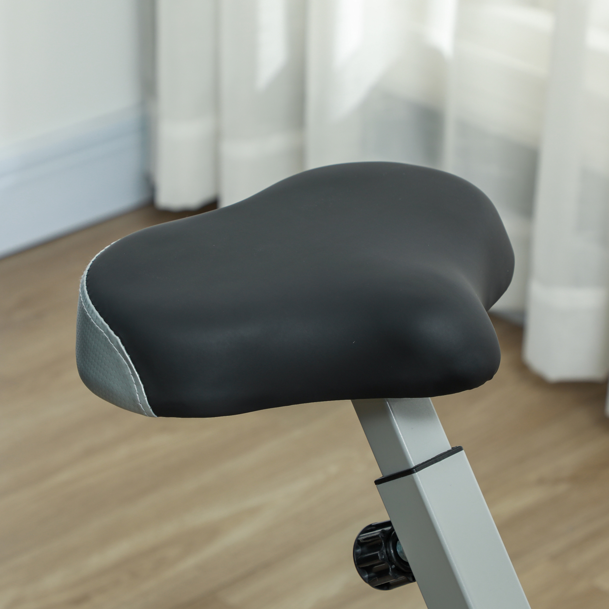 Primo piano di un sellino nero per cyclette. Il sedile è su un palo grigio. Tende e pavimento in legno sullo sfondo.
