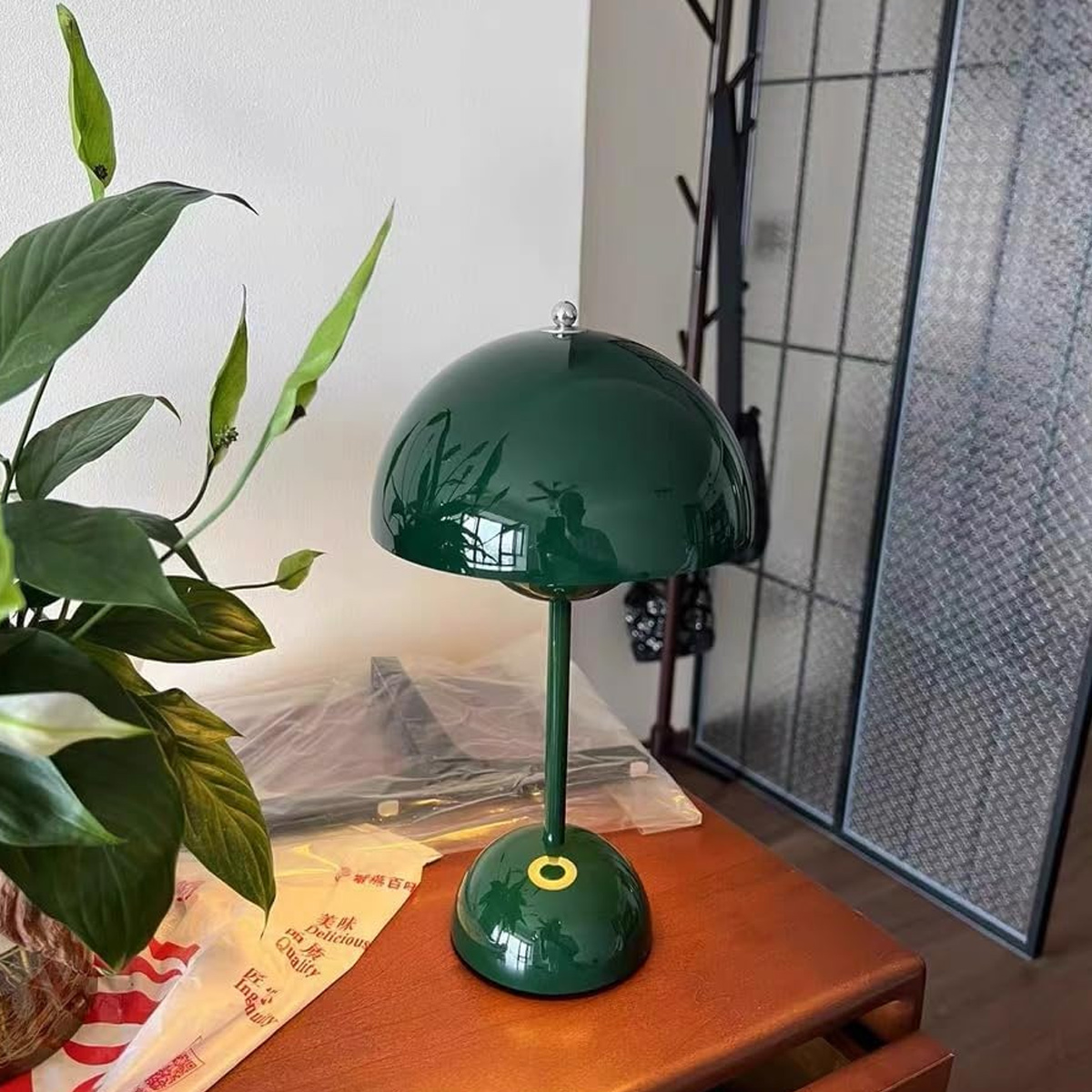 Eine grüne pilzförmige Lampe auf einem Holztisch, links eine Pflanze mit weißen Blüten und im Hintergrund ein dekorativer Schirm.