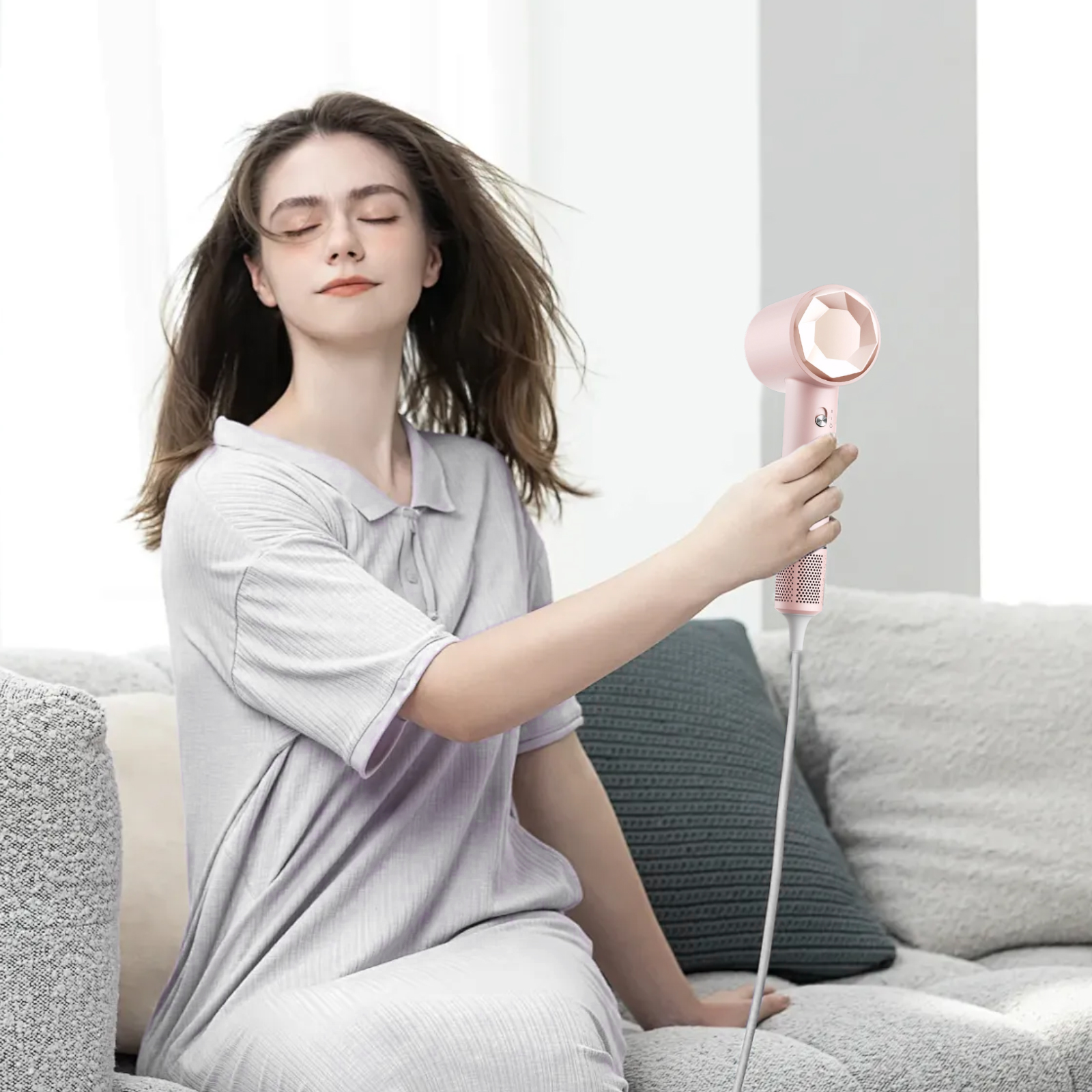 Frau mit geschlossenen Augen, hält einen rosa Haartrockner, sitzt auf einem Sofa. Haare wehen. Hellfarbige Kleidung. Neutraler Hintergrund.