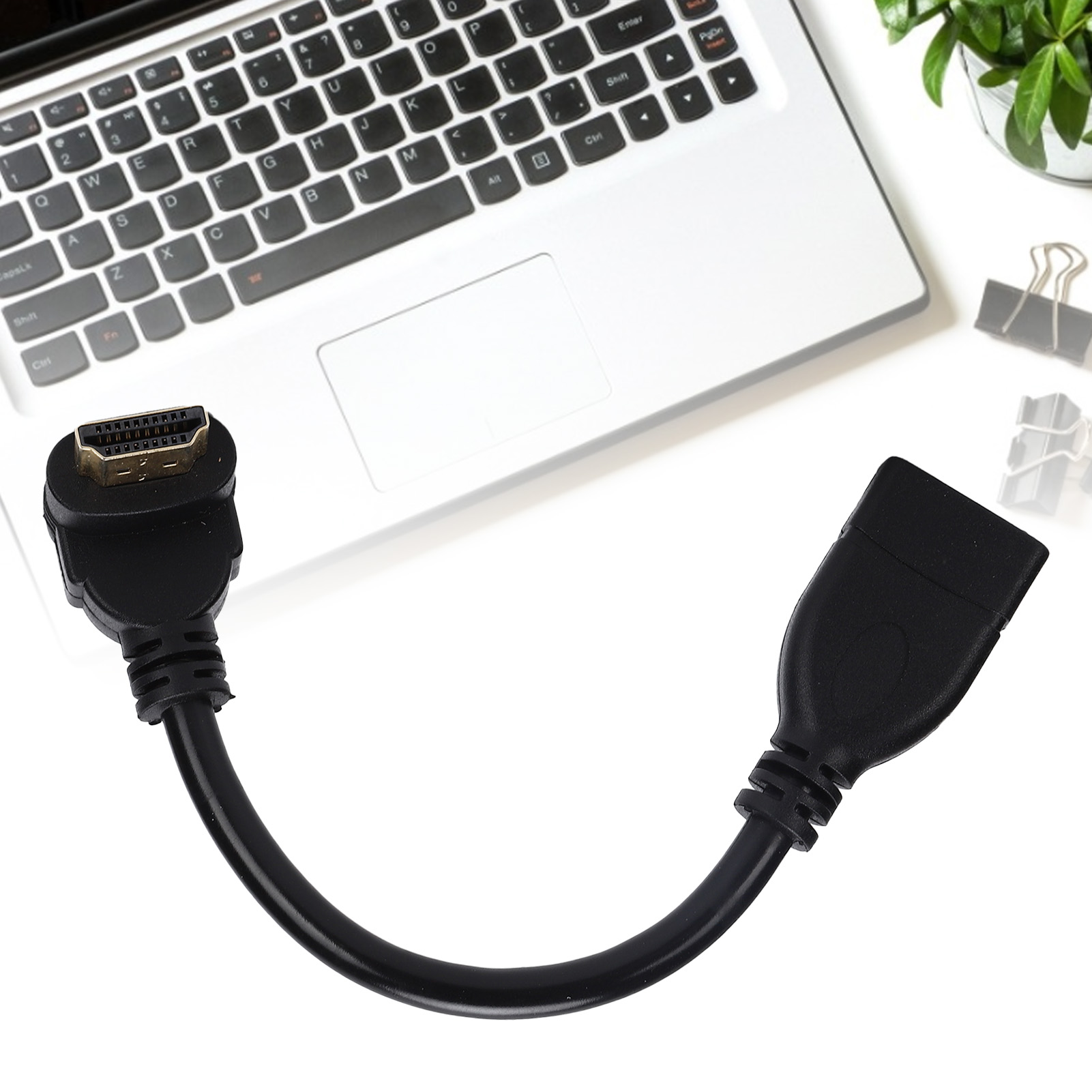 Schwarzes HDMI-Kabel mit Goldstecker, in der Nähe einer Laptop-Tastatur auf weißer Oberfläche. Grüne Pflanze und Büroklammer im Hintergrund.