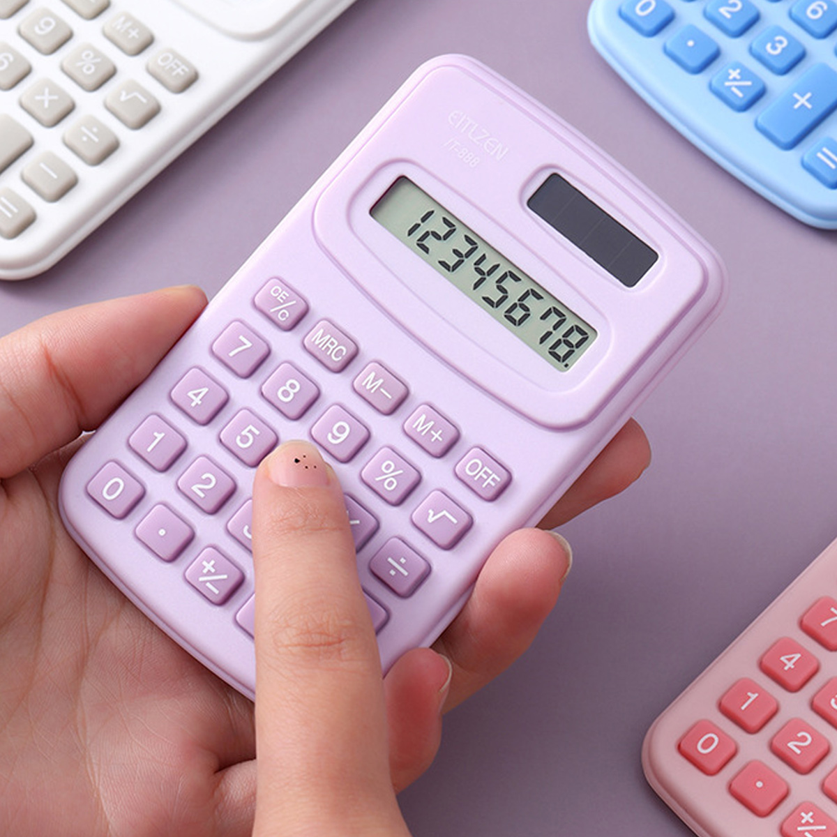 Eine Hand hält einen lavendelfarbenen Taschenrechner mit der Anzeige '12345678'. Weitere Taschenrechner in Weiß, Blau und Rosa sind sichtbar.