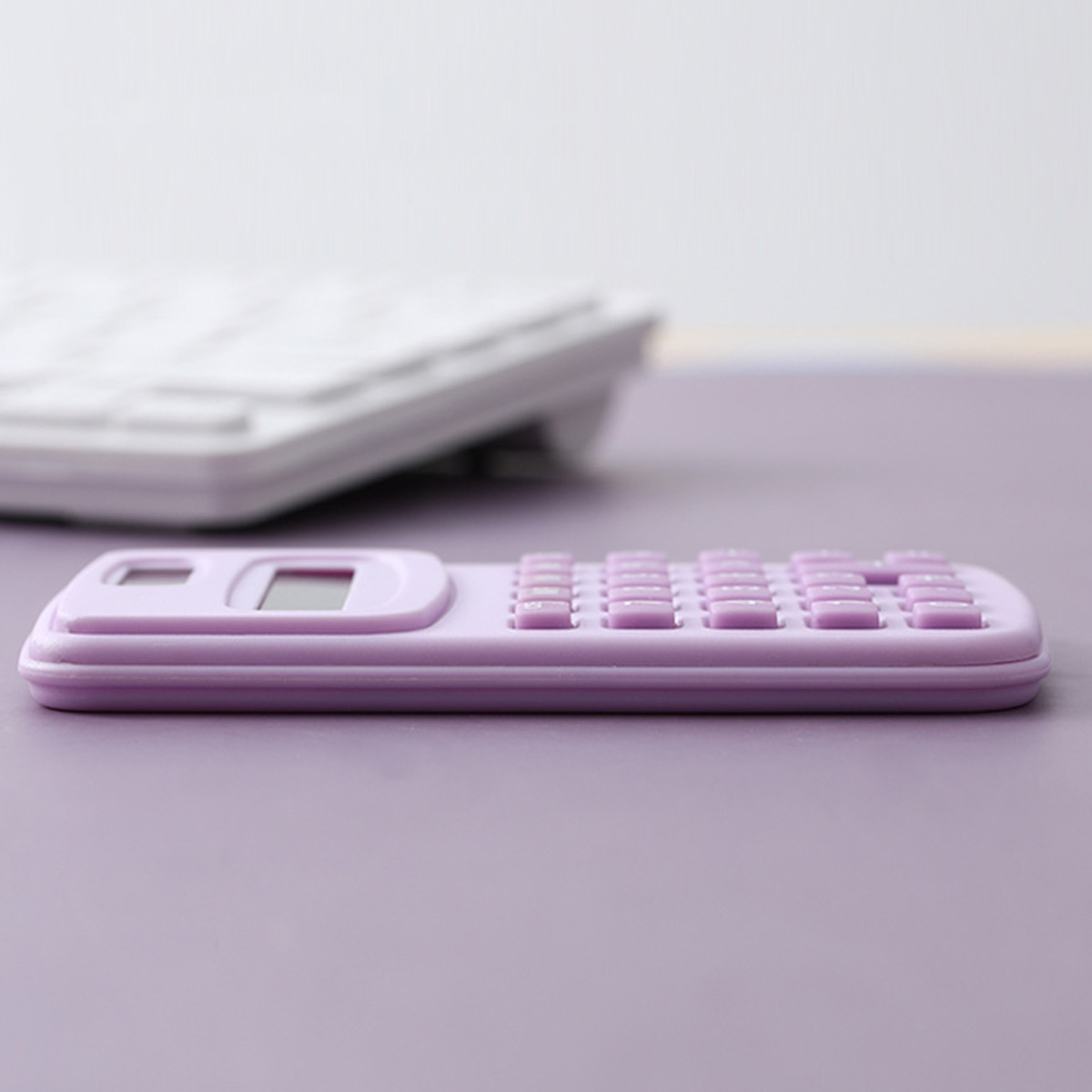 Ein lavendelfarbener Taschenrechner liegt auf einer lilafarbenen Oberfläche. Eine weiße Tastatur ist im Hintergrund unscharf.