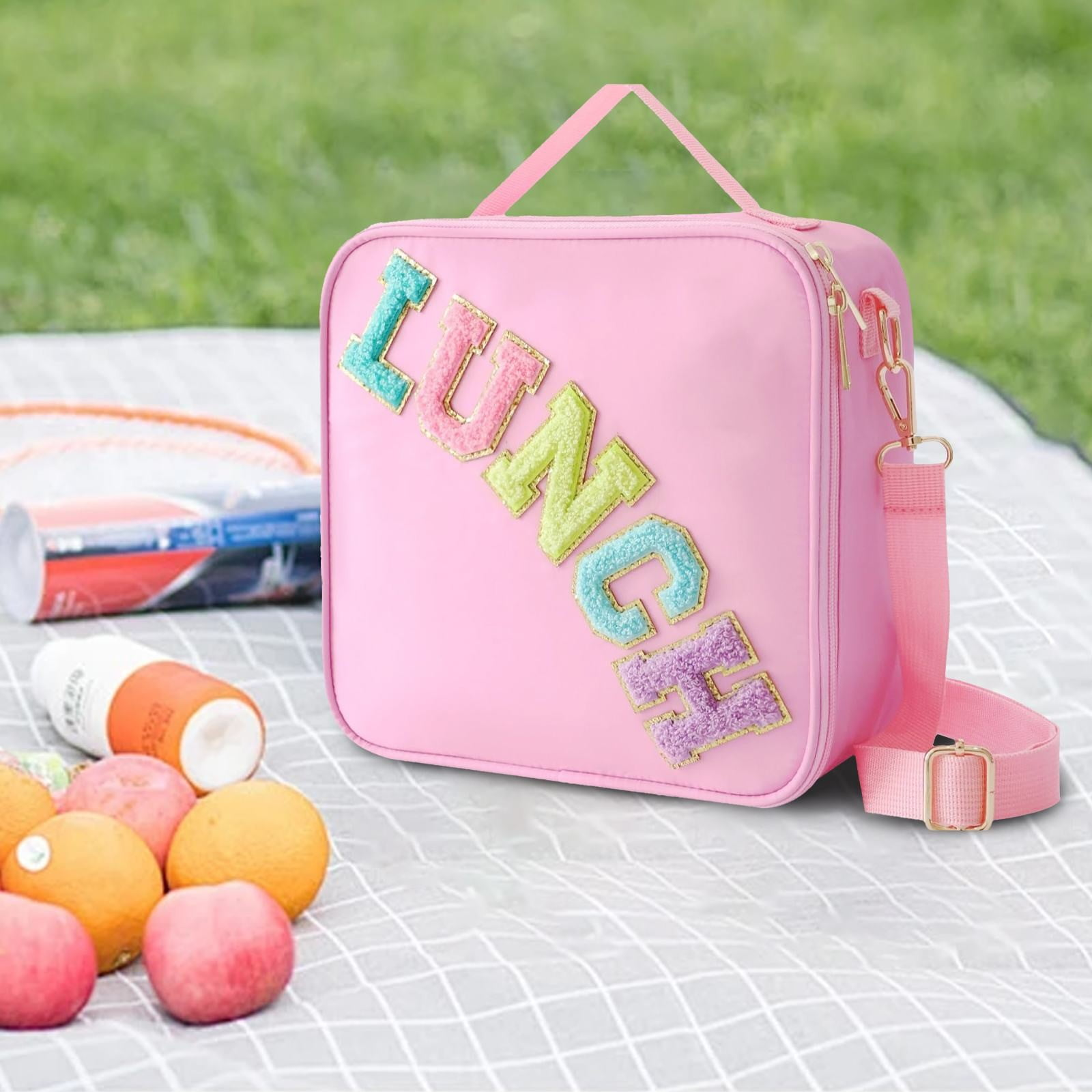 Rosa Lunchpaket mit 'LUNCH'-Text, Obst und Sonnencreme auf einer Picknickdecke. Die Tasche hat einen Schultergurt und einen Griff.