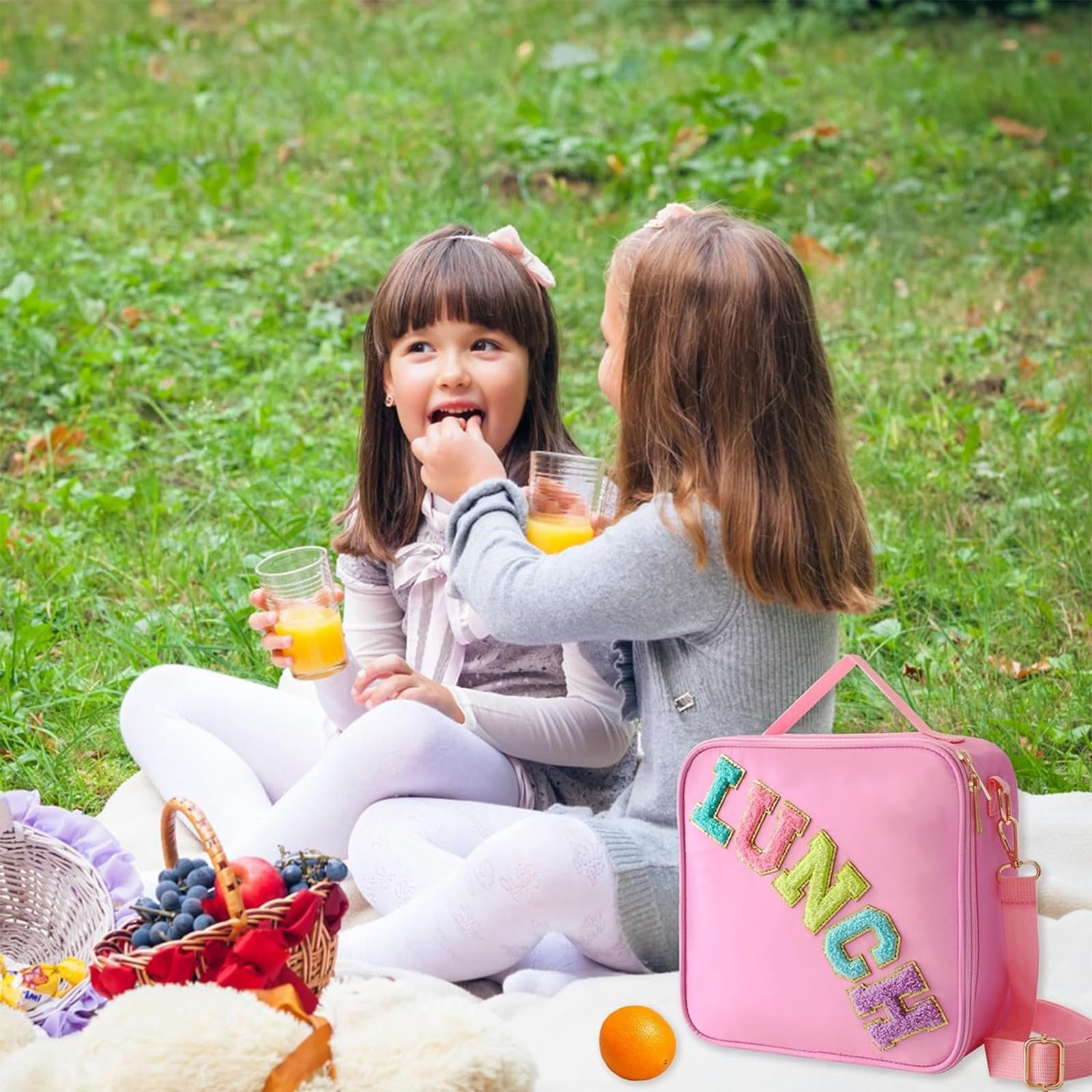 Zwei Mädchen bei einem Picknick. Eines füttert dem anderen Saft. Es gibt eine rosa Lunchtasche, einen Obstkorb und eine Orange auf dem Gras.