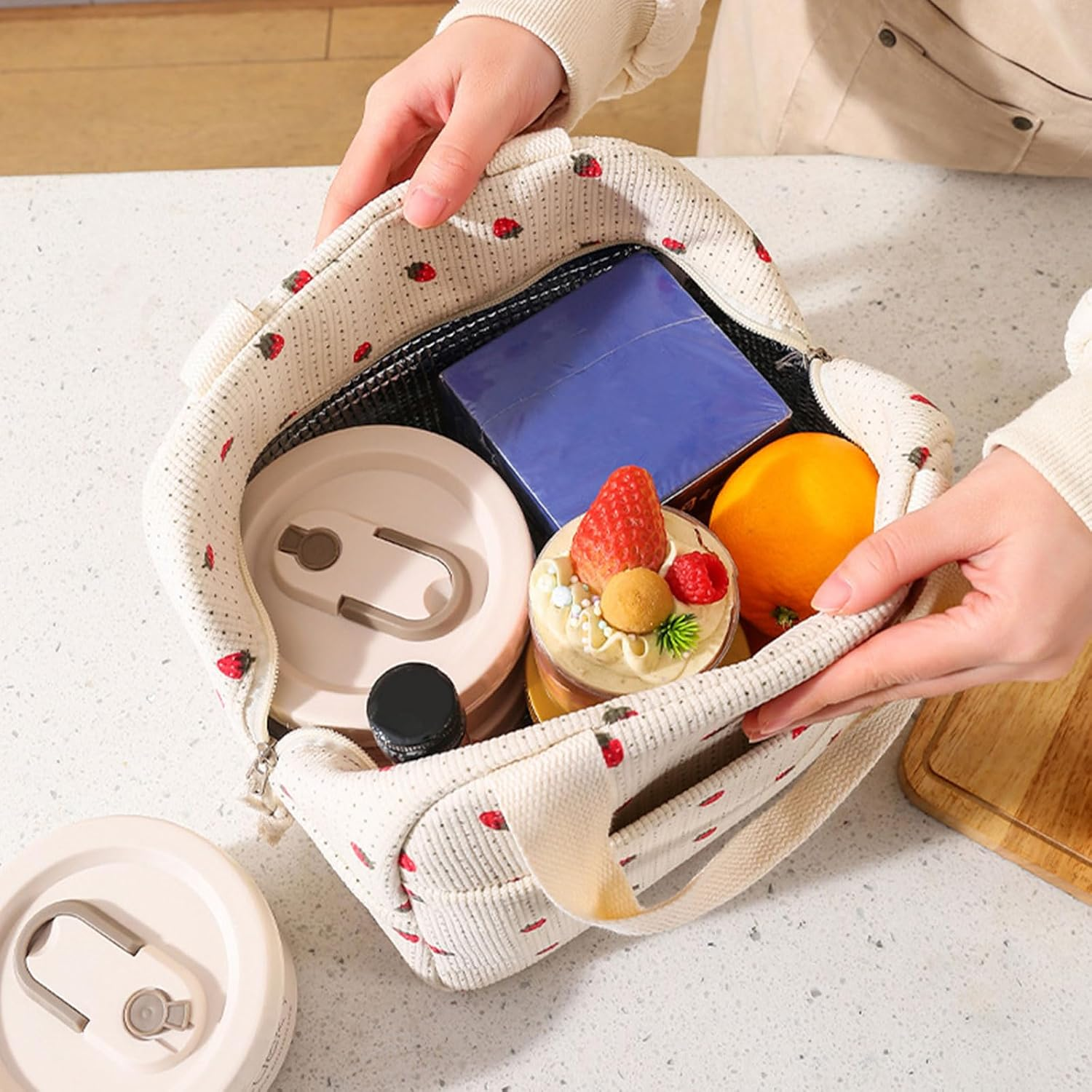 Hände halten eine offene Lunchtasche mit Kuchen, Orange, blauer Box und Behältern. Die Tasche hat Erdbeeren darauf.
