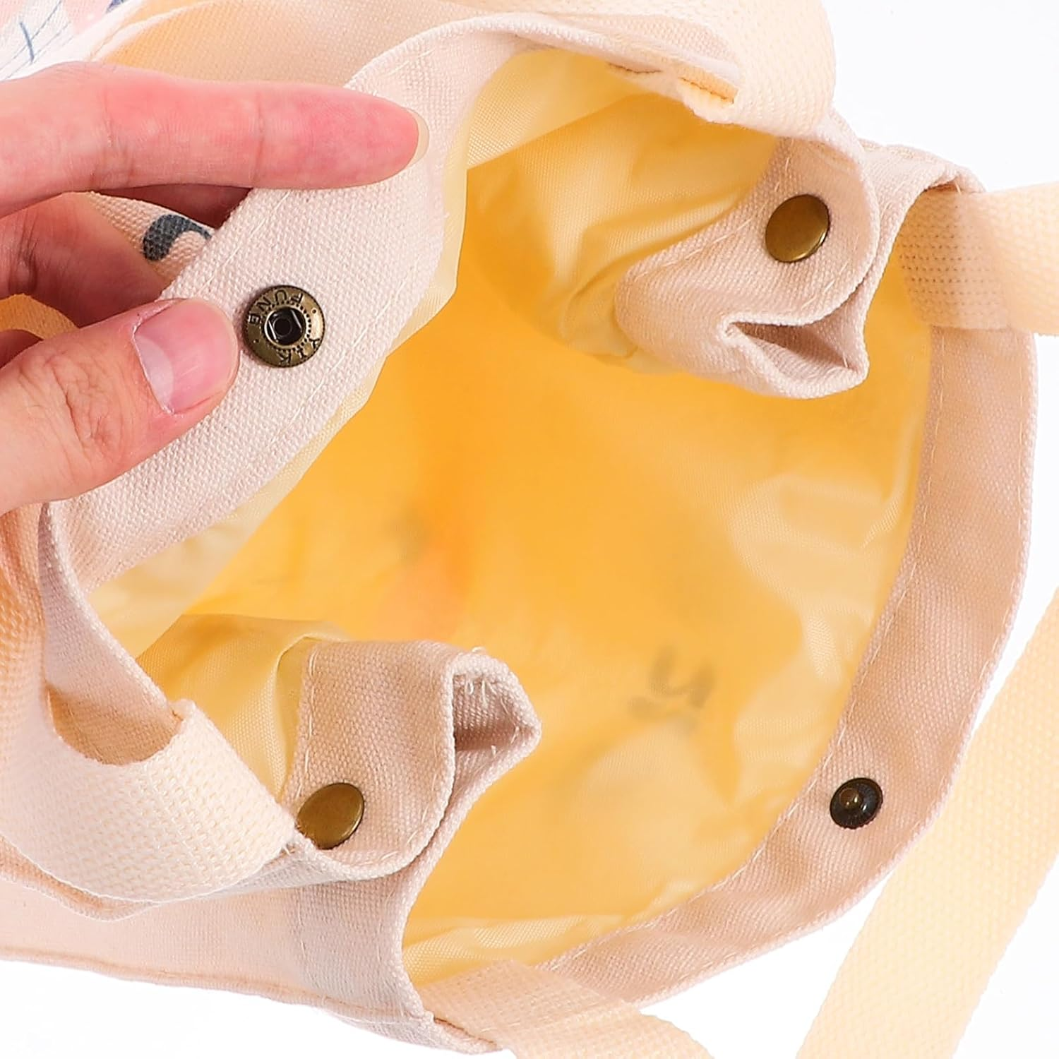 Hand hält eine beigefarbene Tragetasche mit gelbem Futter. Die Tasche hat Druckknöpfe und Schultergurte. Details umfassen Nähte und Metallknöpfe.