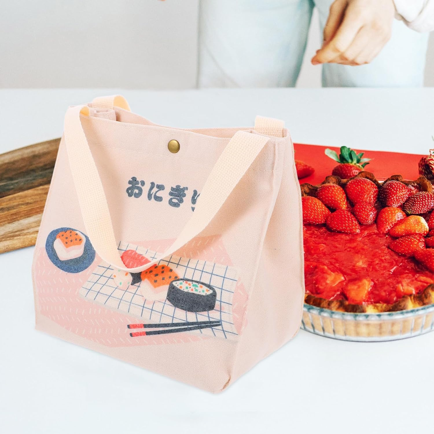 Eine rosa Tasche mit Sushi-Motiv und Griffen steht auf einer weißen Oberfläche. Rechts ist ein Erdbeerkuchen. Japanischer Text ist auf der Tasche.
