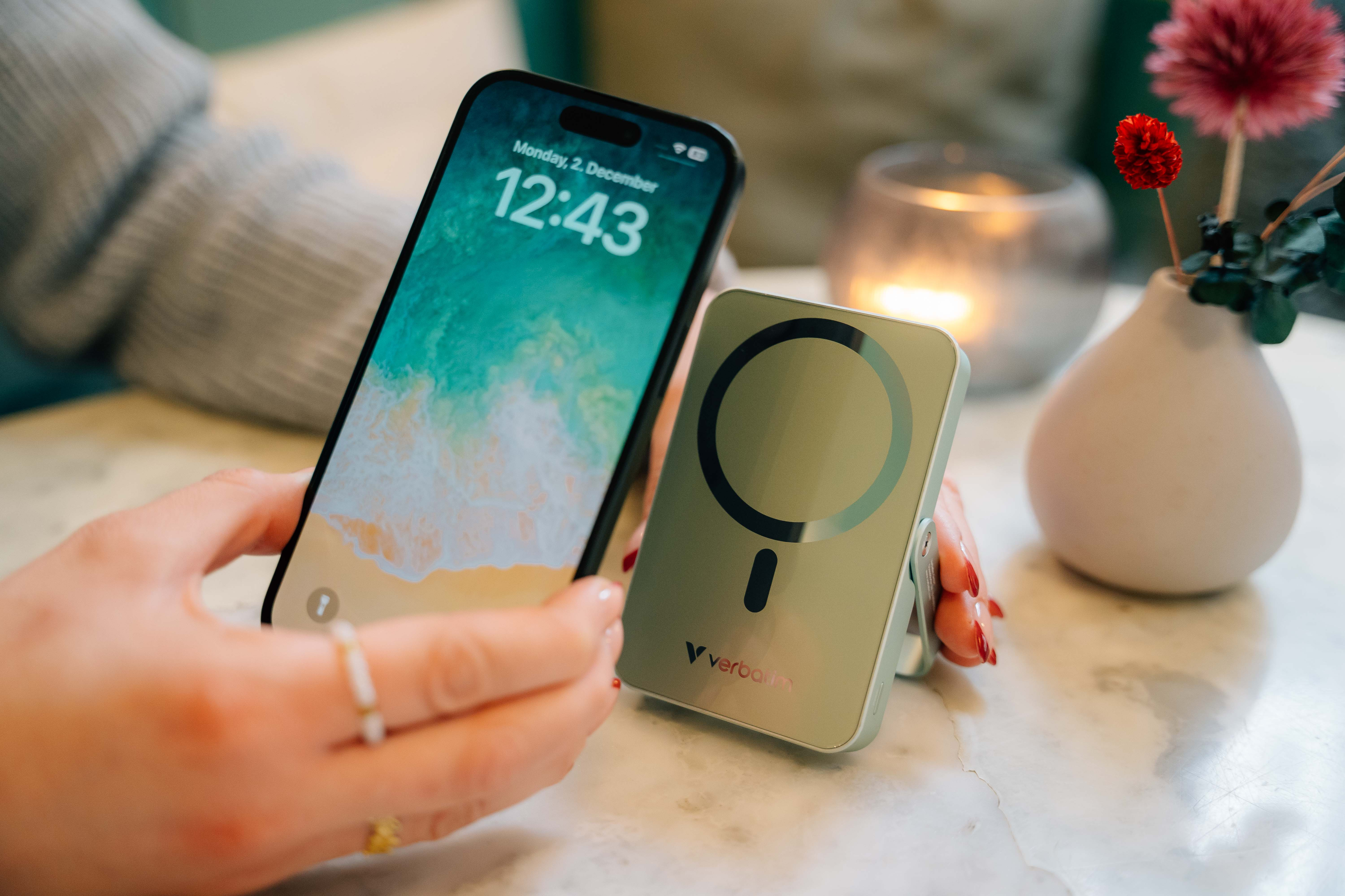 Osoba trzyma telefon i ładowarkę. Ekran pokazuje datę i godzinę. W tle wazon i świeca.