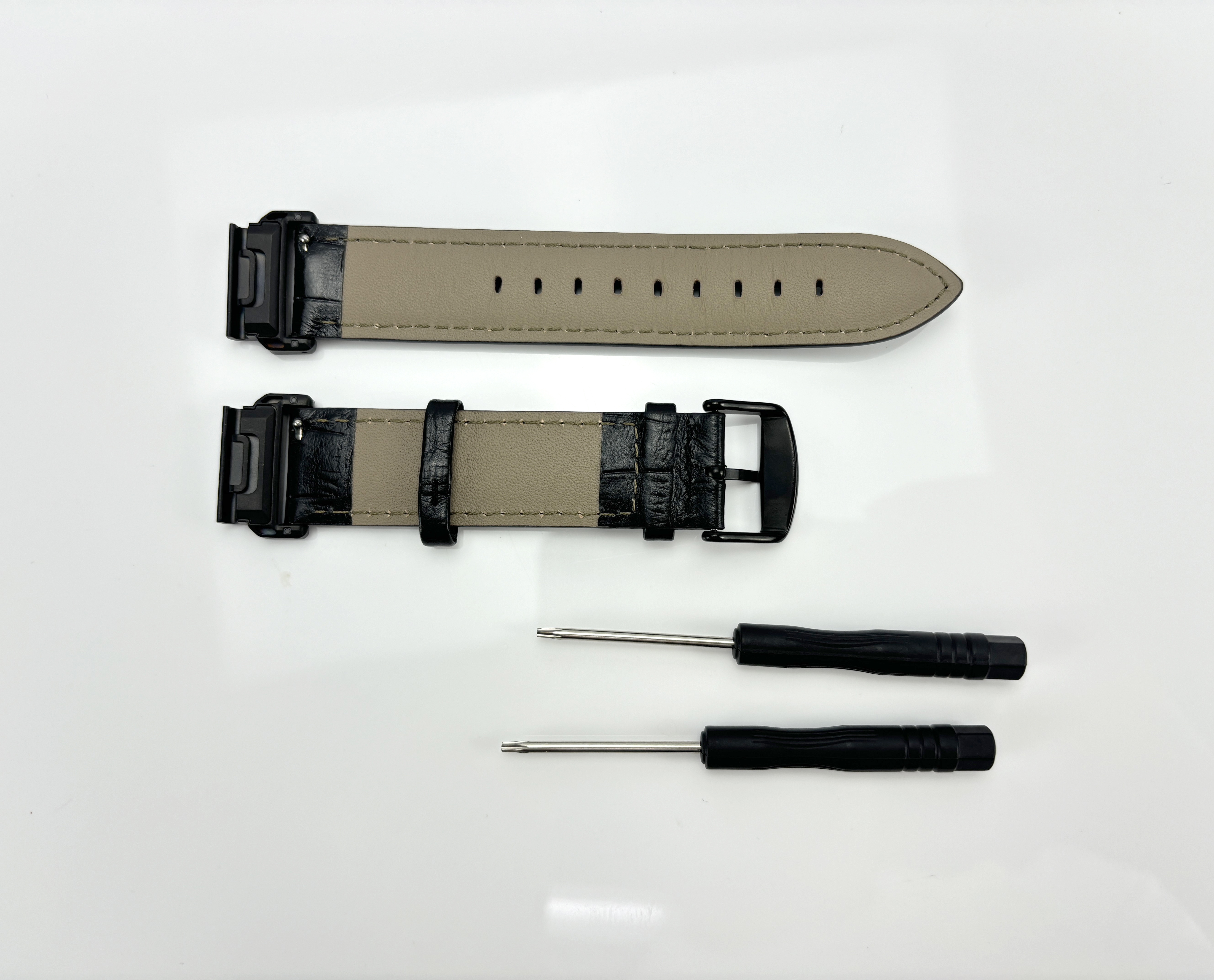 Zwei Uhrenarmbänder und zwei Schraubendreher auf weißer Oberfläche. Armbänder sind schwarz und beige. Schraubendreher haben schwarze Griffe.