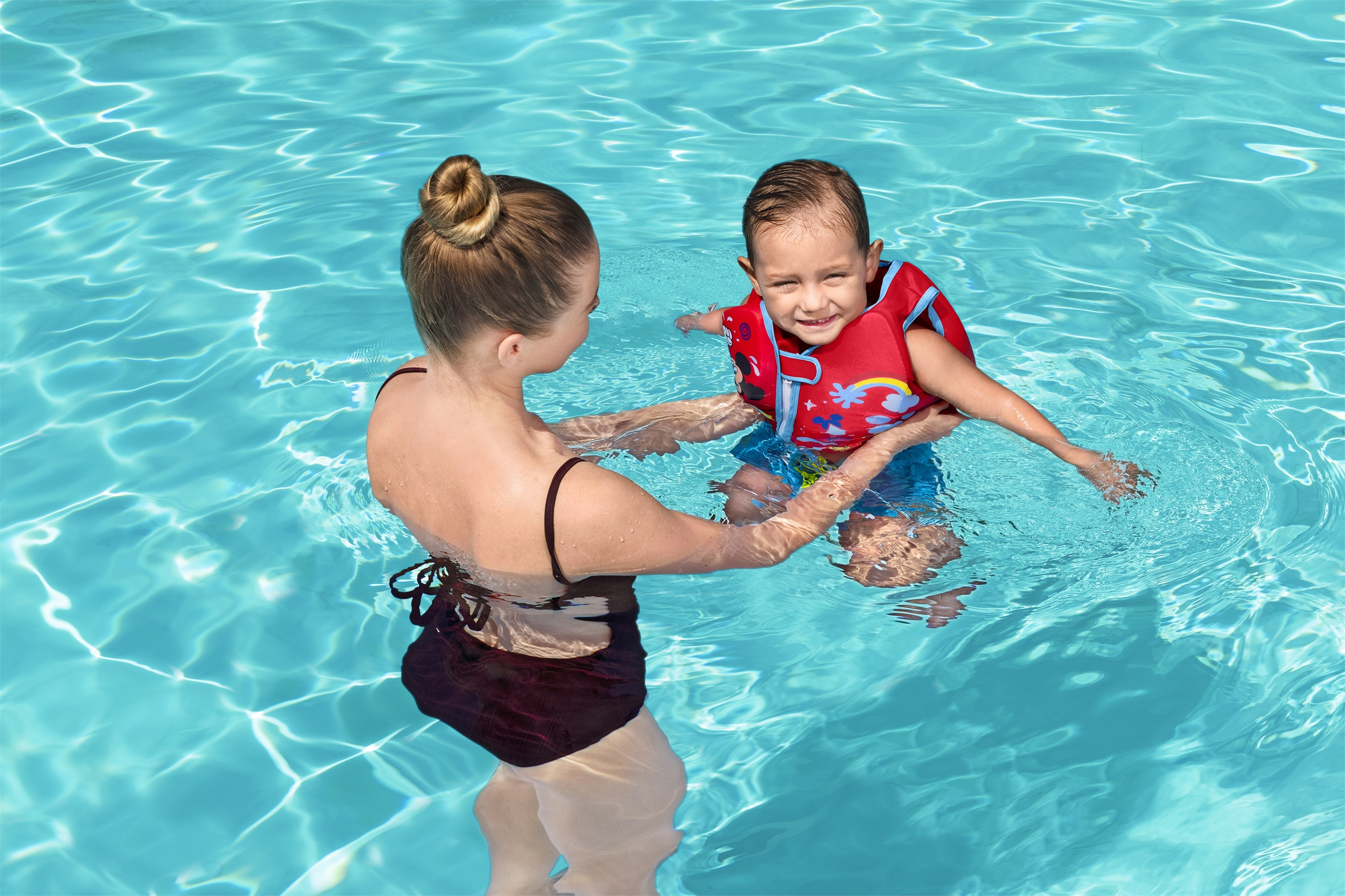 Kobieta trzyma uśmiechnięte dziecko w kamizelce ratunkowej w basenie. Woda jest niebieska.