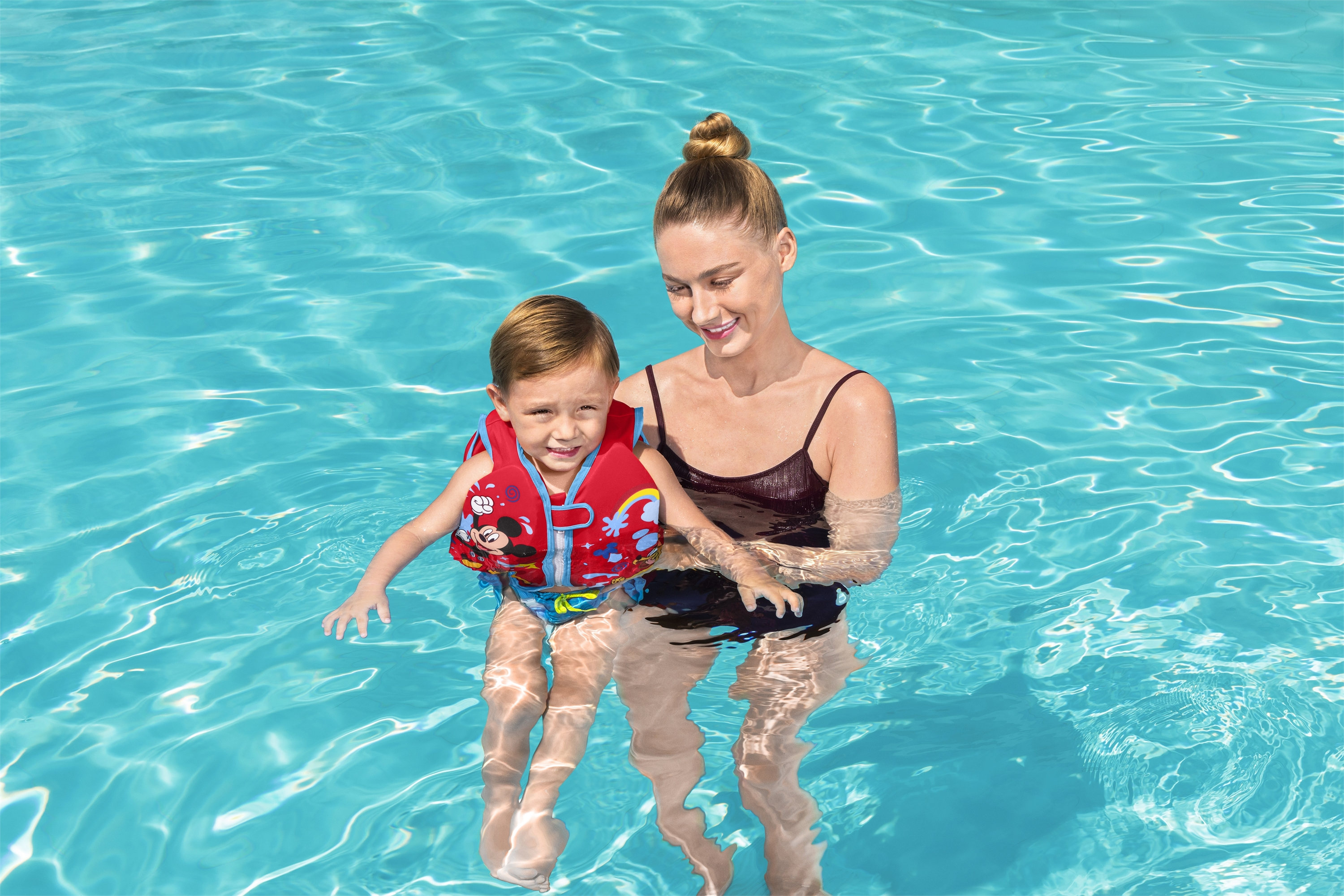 Kobieta trzyma dziecko w basenie. Dziecko ma na sobie czerwoną kamizelkę ratunkową. Oboje się uśmiechają.