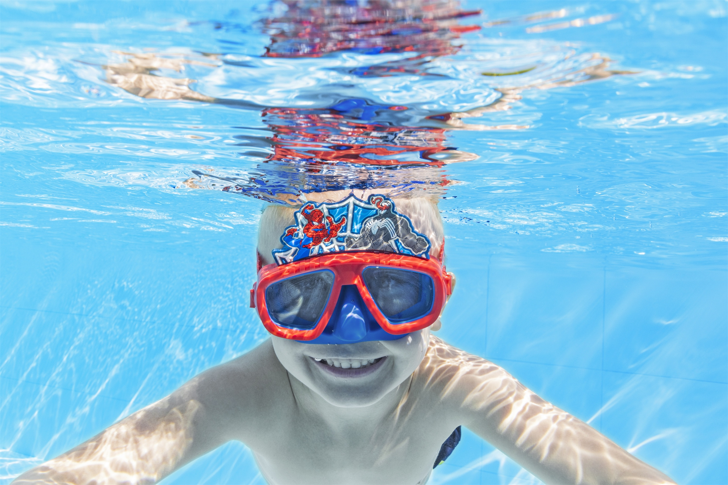 Un bambino sott'acqua, con occhialini rossi e blu con un design da supereroe, sorridente. L'acqua blu riempie la scena.