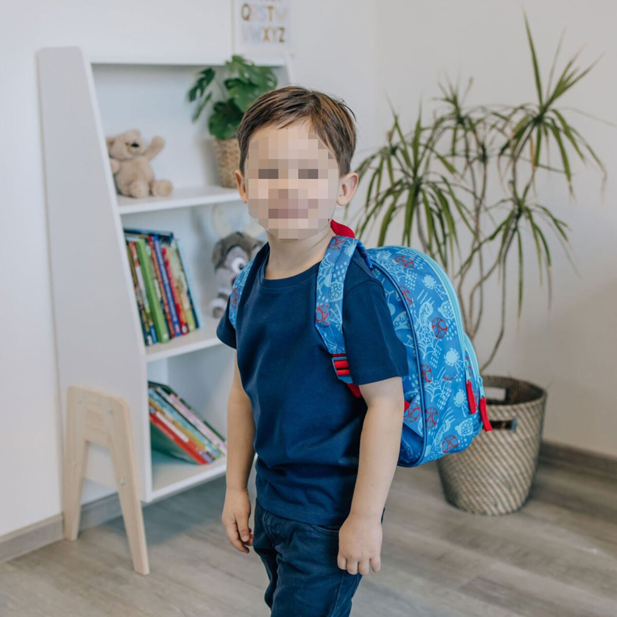 A young boy with a blue backpack stands in front of a white shelf. A plant and a basket are in the background.