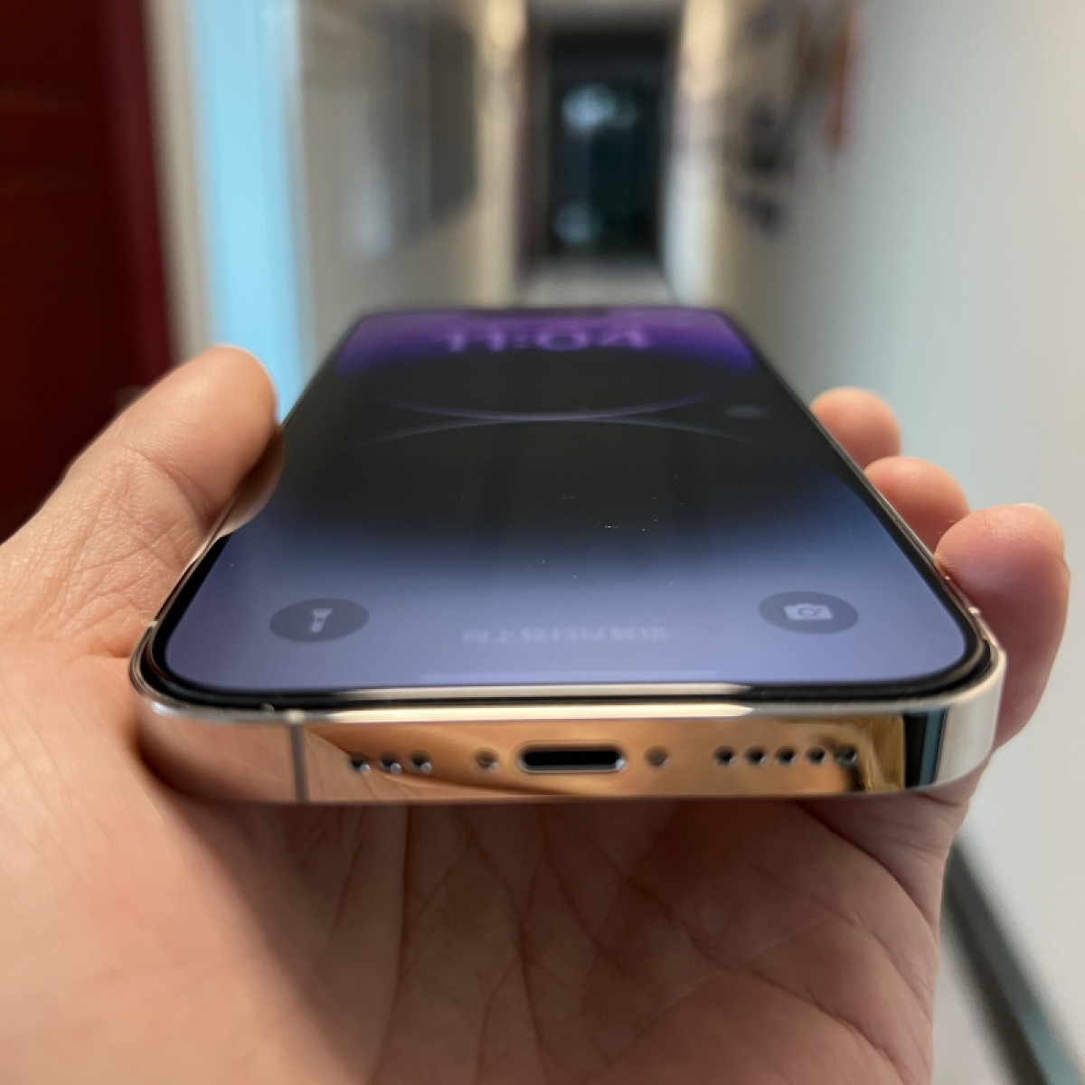 Una mano che tiene un telefono. Lo schermo mostra una schermata di blocco. Il telefono è argento, con uno schermo nero. Lo sfondo è un corridoio.