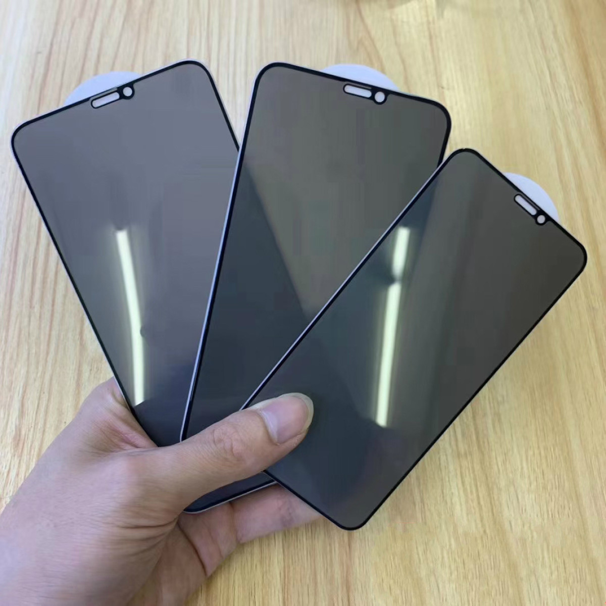 Una mano tiene tre protezioni per lo schermo dello smartphone. Sono grigie scure, rettangolari e hanno un ritaglio per la fotocamera. Lo sfondo è in legno.