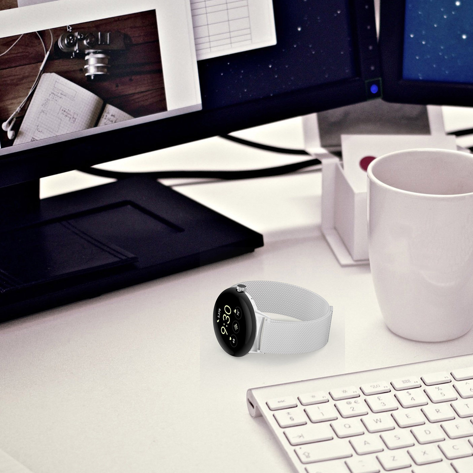 Uno smartwatch con cinturino bianco è appoggiato su una scrivania bianca. Sono presenti anche una tastiera, una tazza e monitor del computer.