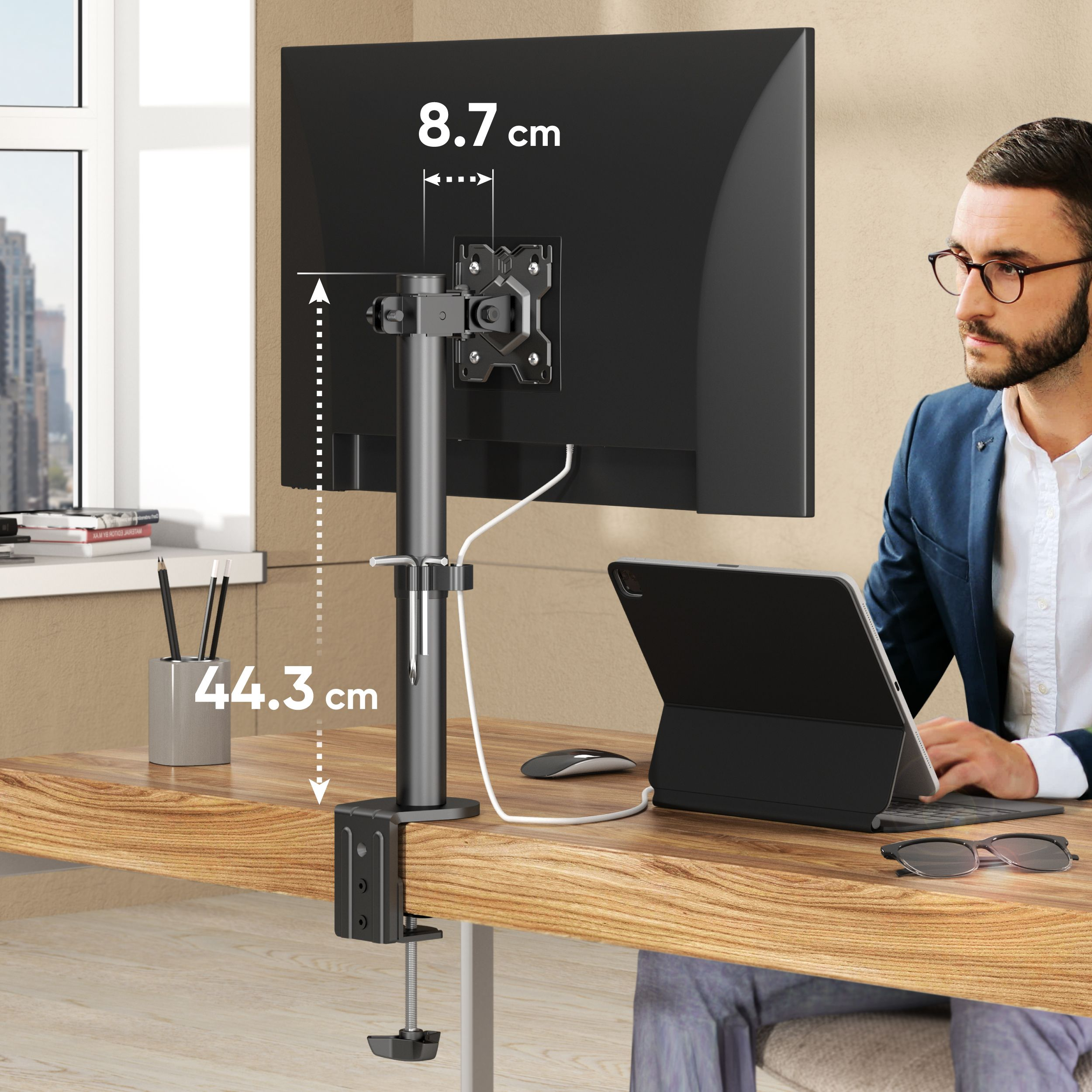 Un hombre trabaja en una tableta en un escritorio. Un soporte de monitor sostiene una pantalla. Se indican las medidas.