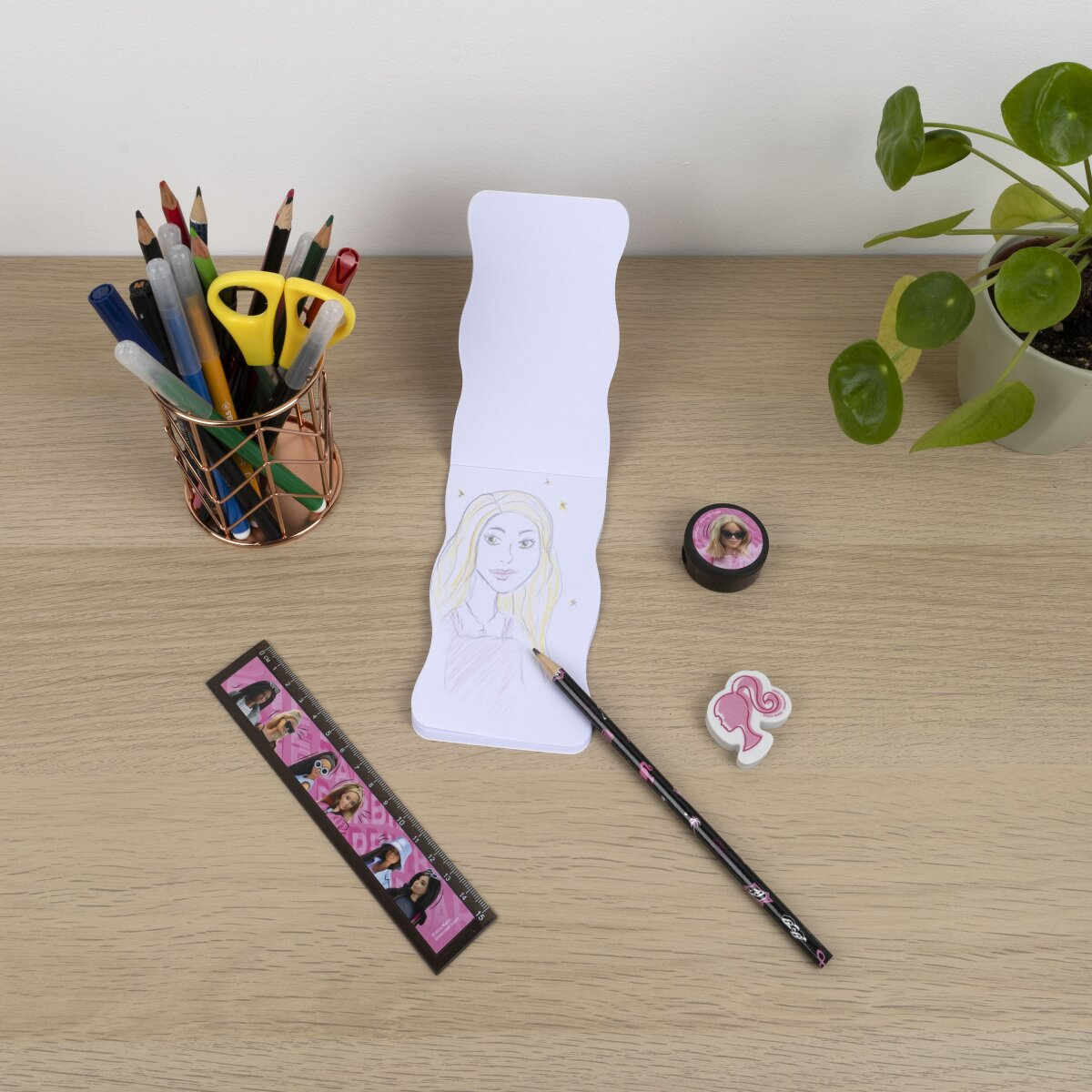 A desk with a drawing, pencils, a ruler, a sharpener, a Barbie eraser, and a plant in the background.