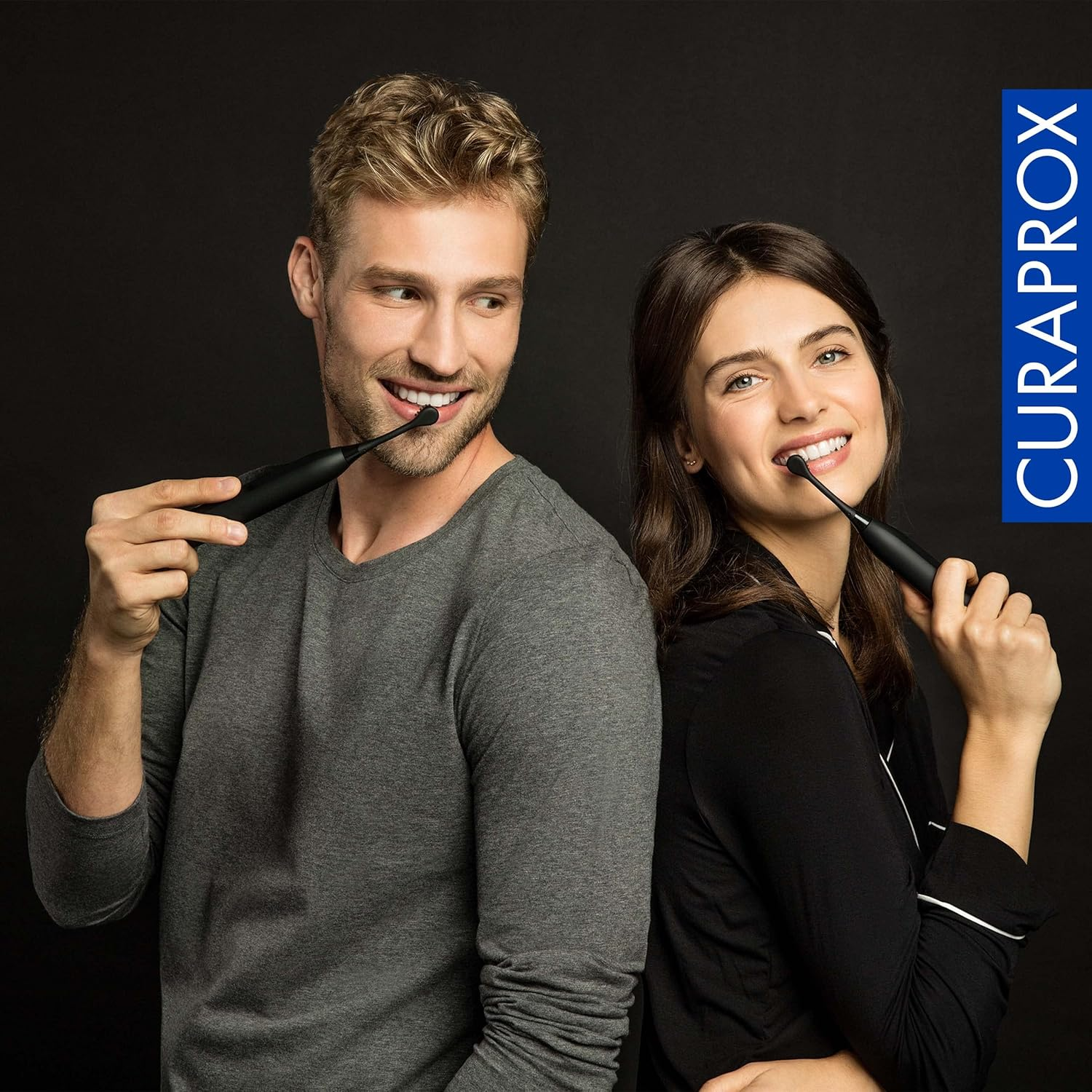 Mężczyzna i kobieta trzymają czarne szczoteczki do zębów, uśmiechając się. Kobieta ma na sobie czarną bluzkę, mężczyzna szarą. Logo Curaprox po prawej.