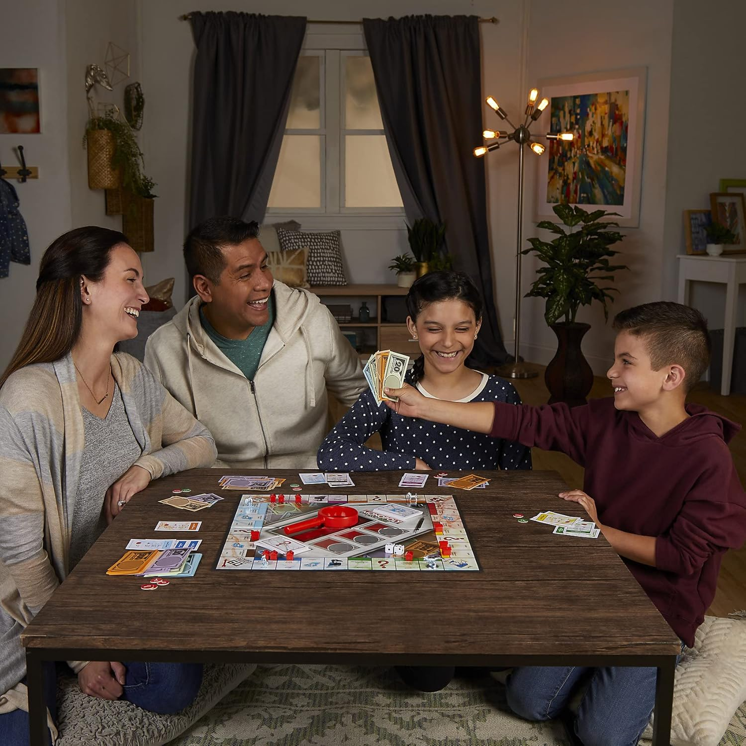 Rodzina gra w Monopoly. Cztery osoby wokół drewnianego stołu z grą planszową. Śmieją się i przekazują pieniądze.