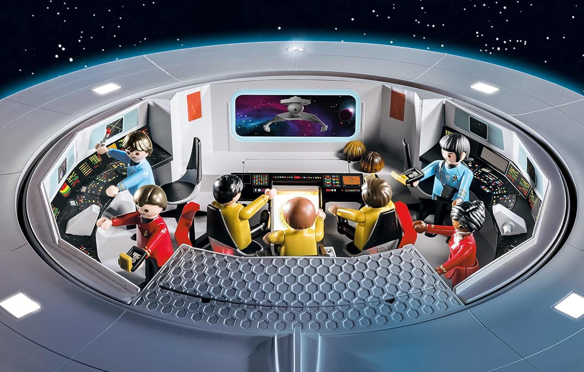 Una scena del ponte giocattolo di Star Trek. Figure in uniforme sedute alle console, che guardano gli schermi. Una scena spaziale è visibile su un monitor centrale.