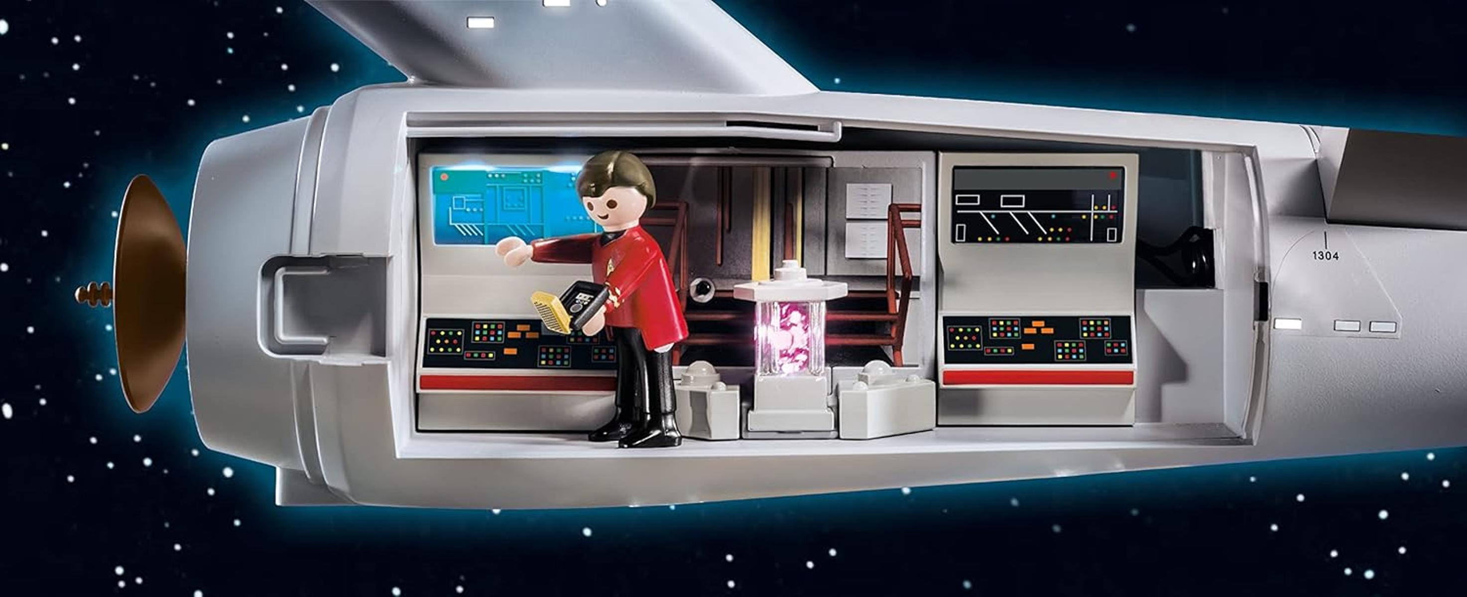 Interno dell'astronave giocattolo di Star Trek con una figura in uniforme rossa, che tiene in mano un dispositivo. Pannelli di controllo e sfondo spaziale.