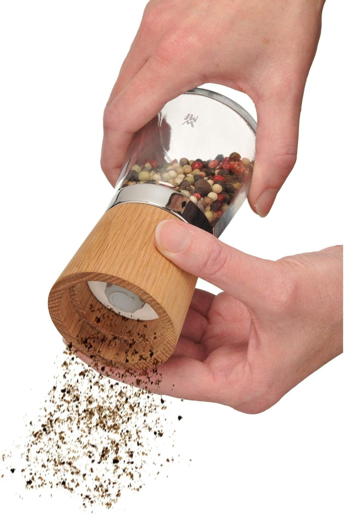 Eine Person mahlt Pfeffer mit einer Pfeffermühle aus Holz und Glas. Pfeffer fällt heraus. Die Mühle wird mit beiden Händen gehalten.
