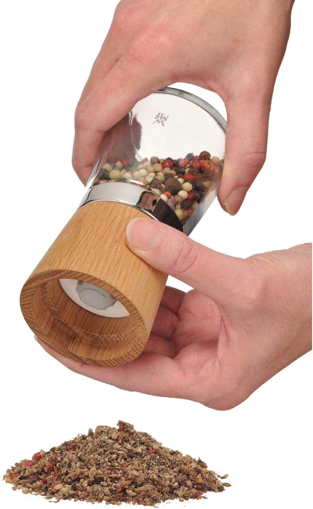 Hände halten eine Pfeffermühle und mahlen Pfefferkörner in einen Haufen gemahlenen Pfeffers. Die Mühle ist aus Holz und Glas.