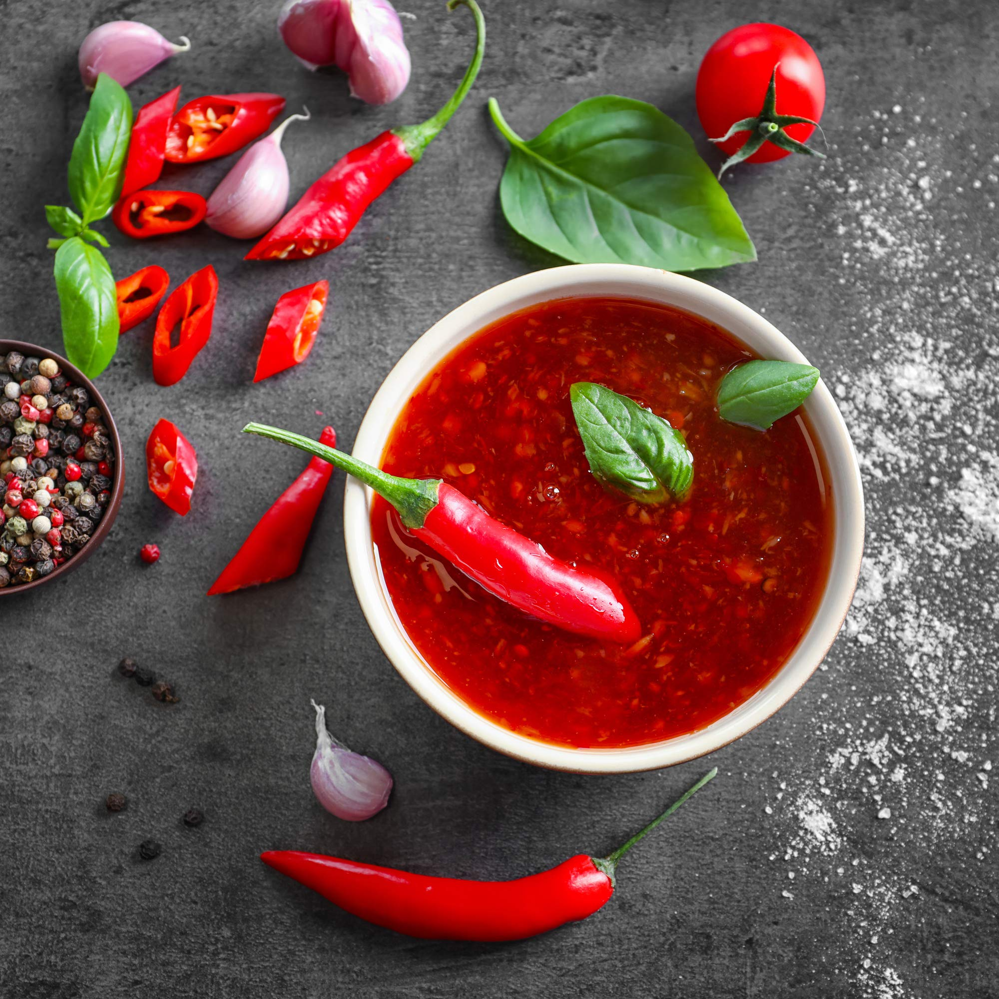 Un bol de sauce rouge entouré de piments, d'ail, de basilic, de grains de poivre et d'une tomate sur une surface grise foncée.