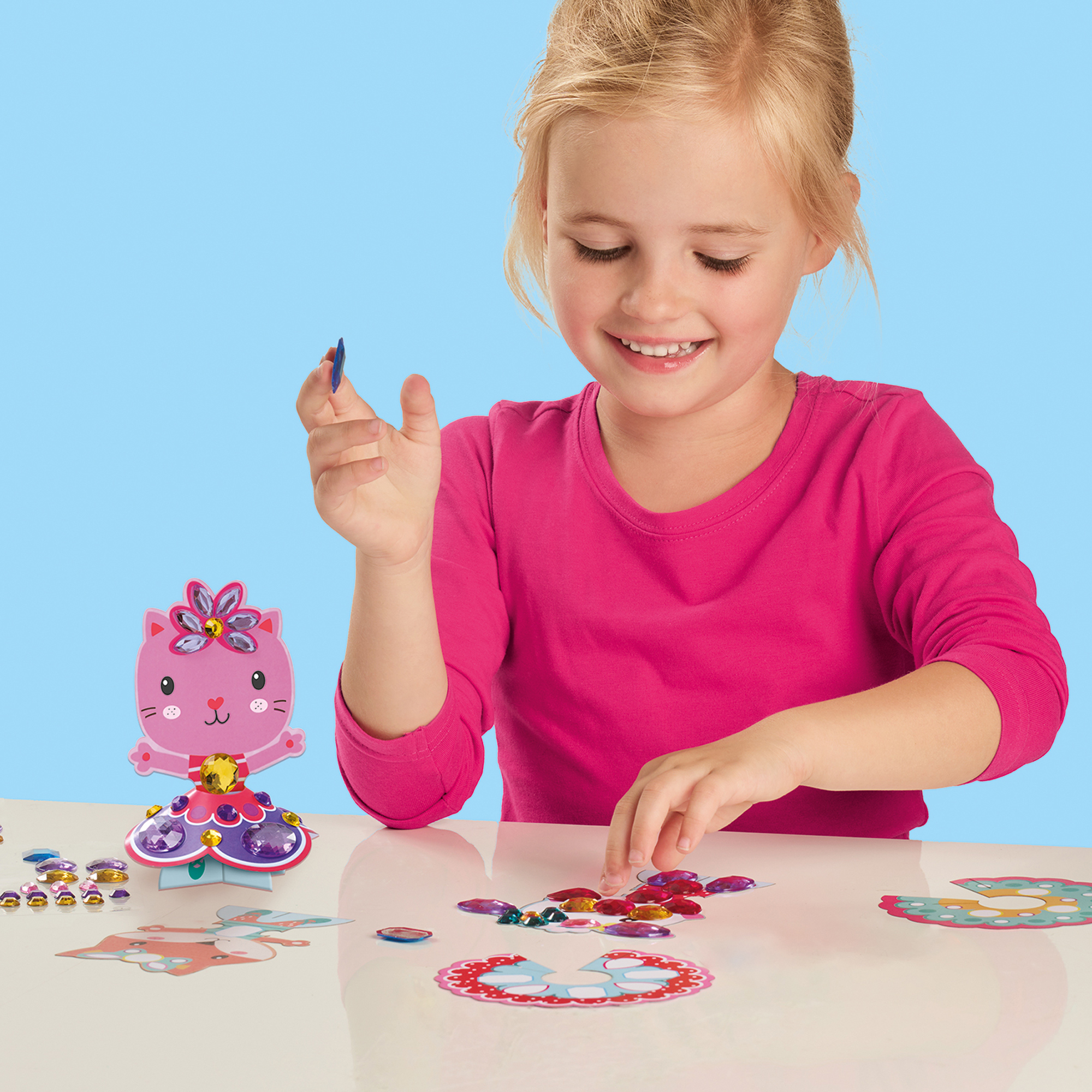 Dziewczyna uśmiecha się podczas tworzenia. Na stole figurka różowego kota i klejnoty. Trzyma niebieską kredkę.