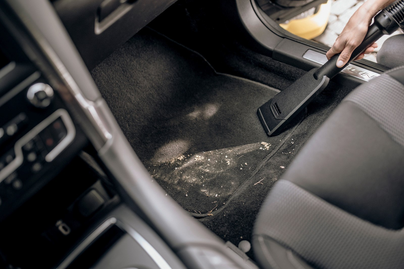 Una persona aspira l'interno di un'auto. La testina nera dell'aspirapolvere è sul tappetino sporco. Il sedile dell'auto è a destra.