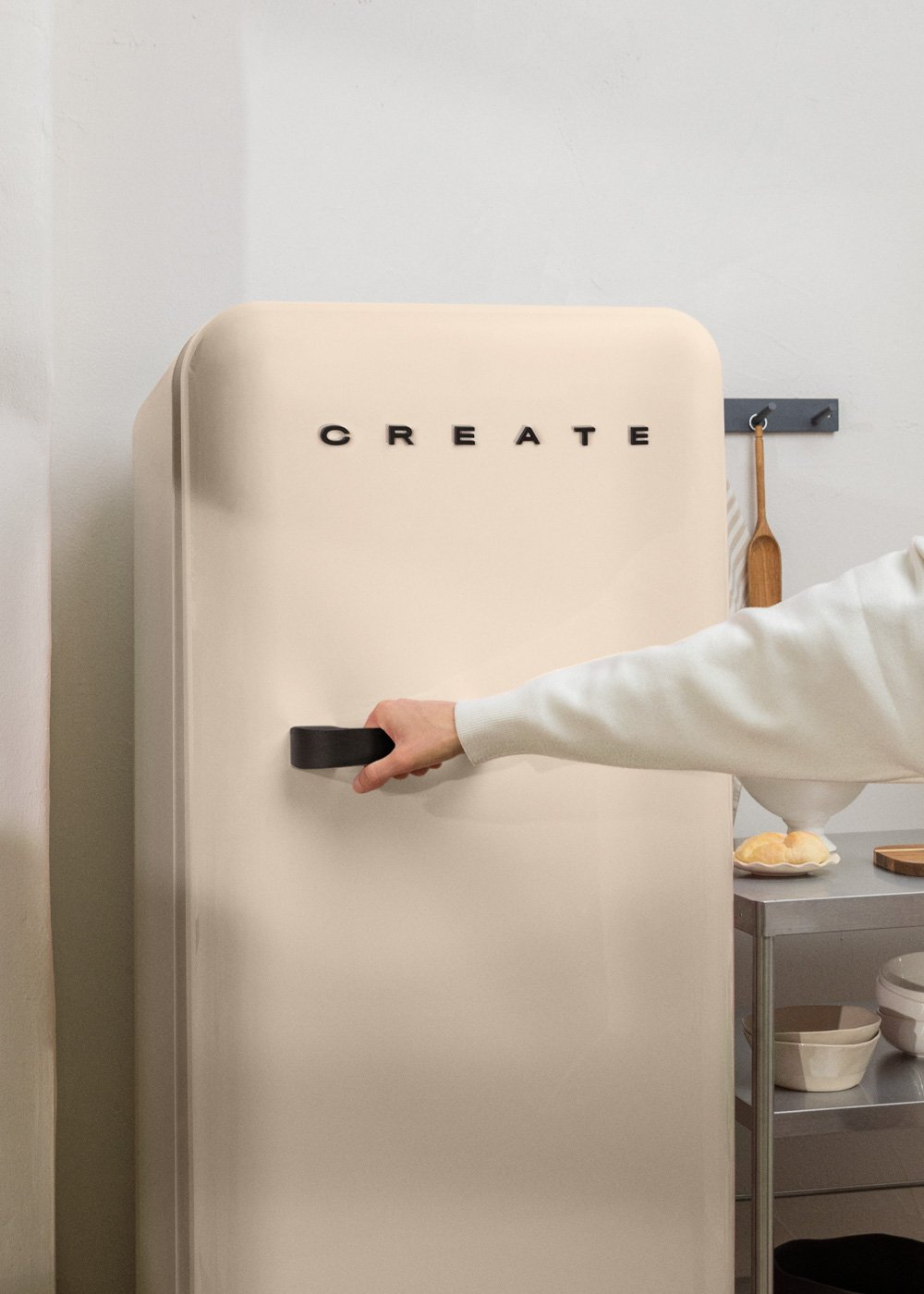 Una persona abre una nevera beige con 'CREATE' en ella. Cerca hay una cuchara de madera y cuencos. La escena es una cocina.