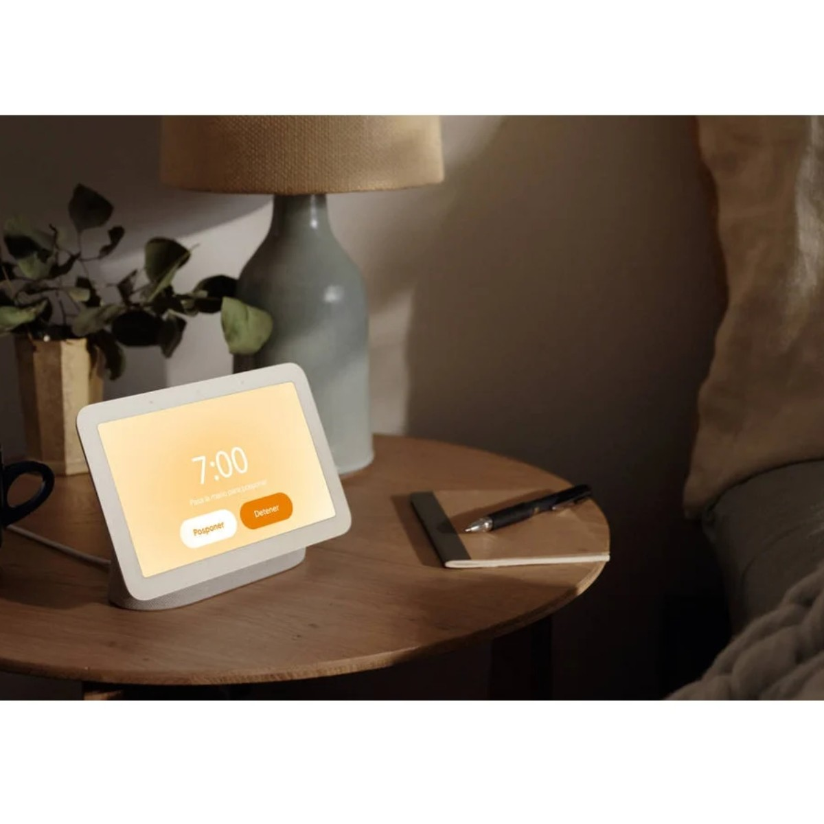 Un display intelligente mostra le 7:00 su un comodino. Accanto, una lampada, un taccuino e una penna. Una pianta e un letto sullo sfondo.