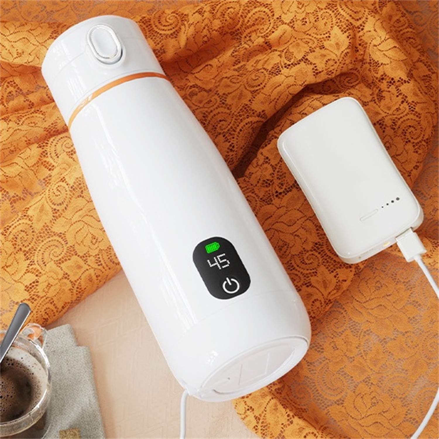 Weißer Reisebecher mit Digitalanzeige neben einer weißen Powerbank. Beide auf orangefarbenem Spitzengewebe. Eine Tasse Kaffee ist ebenfalls zu sehen.