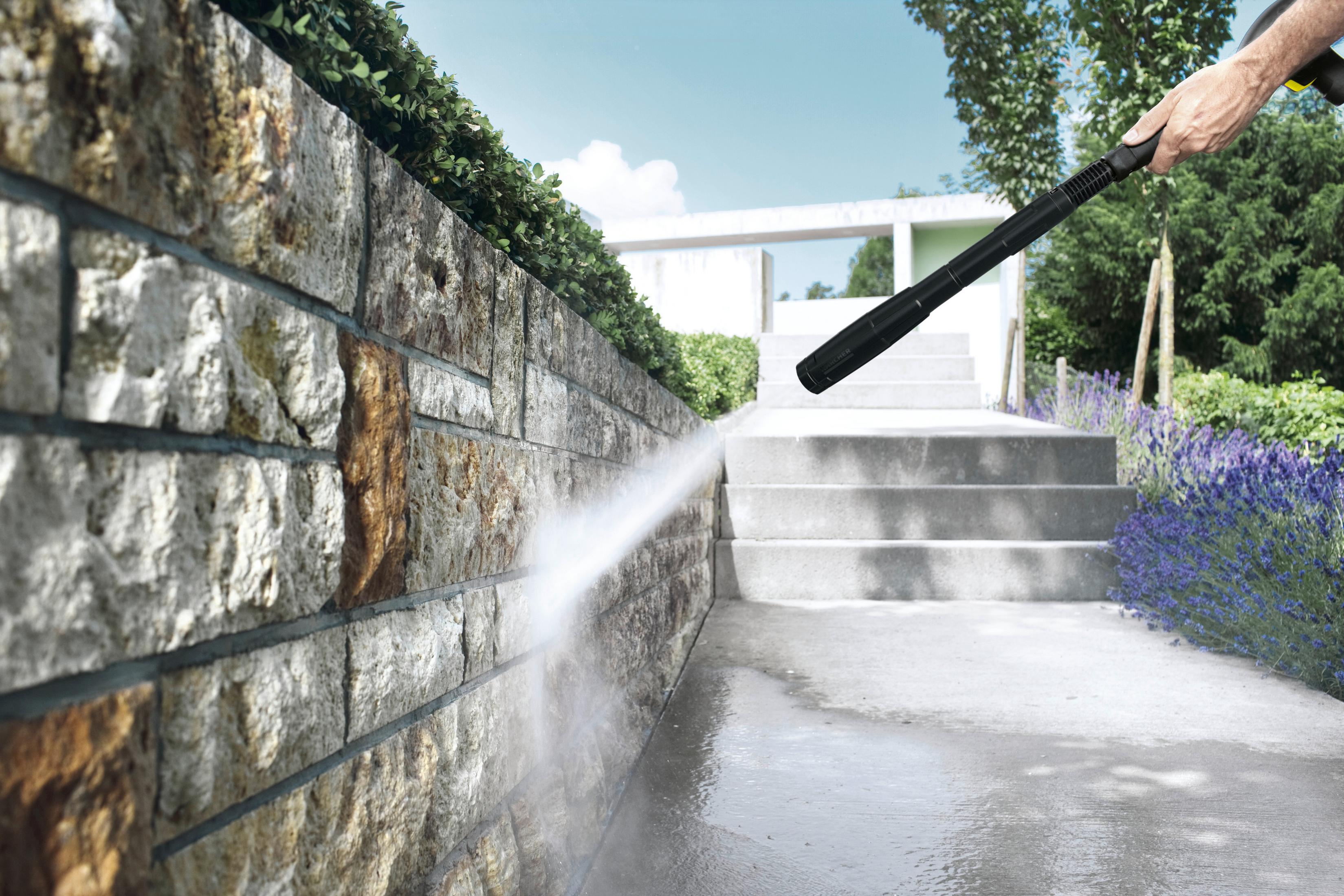 Osoba myje ciśnieniowo kamienny mur. Woda tryska z czarnej dyszy. W tle schody i budynek.