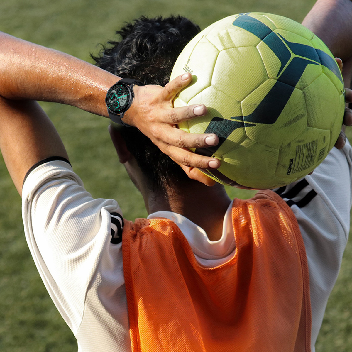 Ein Fußballspieler hält einen gelben Fußball über seinem Kopf. Er trägt eine orangefarbene Weste und eine Smartwatch. Der Hintergrund ist grünes Gras.