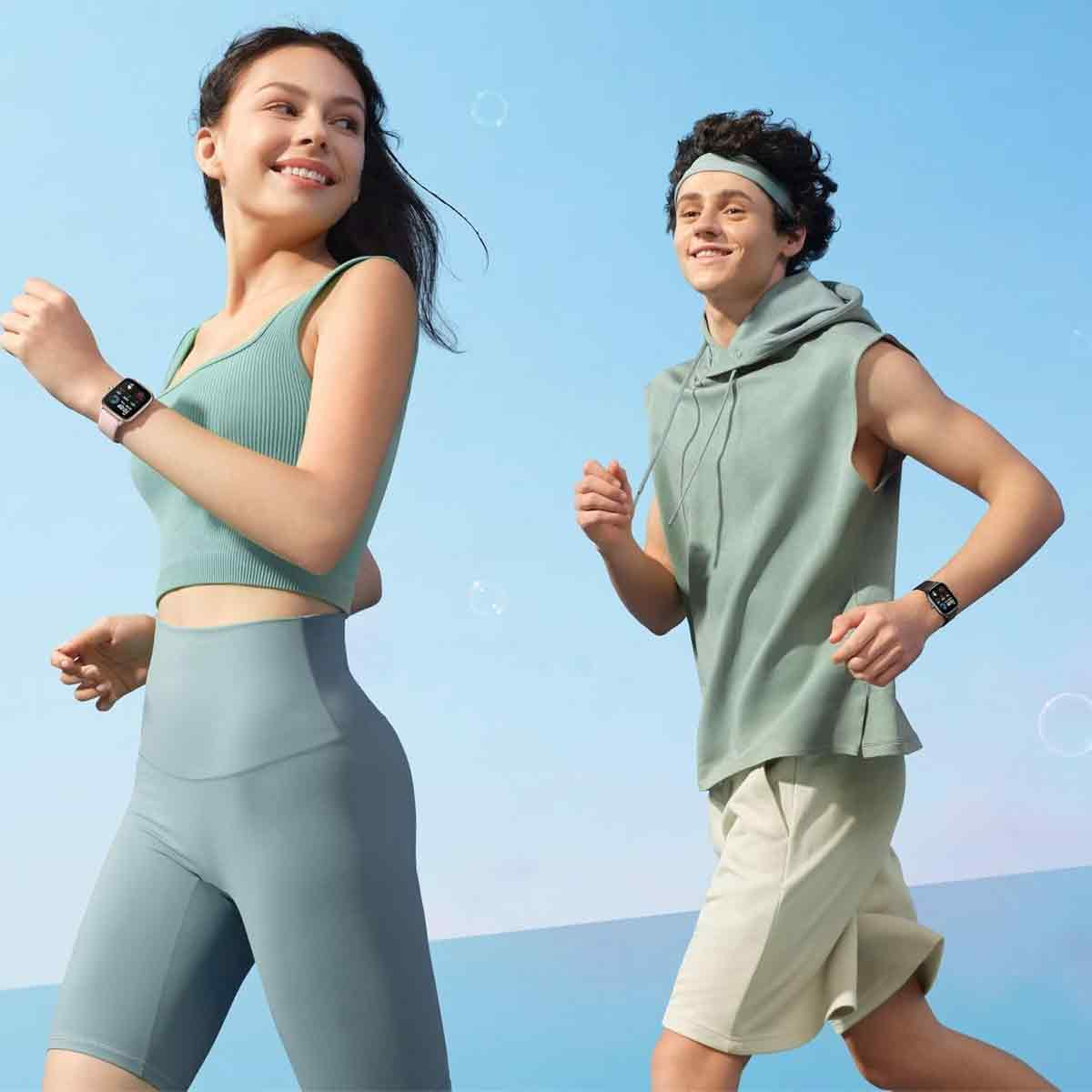 Zwei Personen beim Laufen. Frau in grünem Top und Shorts, Mann in ärmellosem Kapuzenpullover. Beide tragen Smartwatches. Blauer Hintergrund.