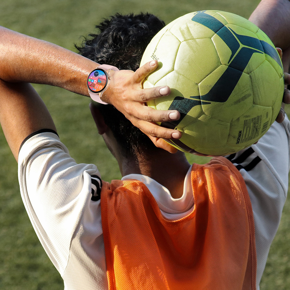 Ein Fußballspieler hält einen gelben Ball über seinen Kopf. Er trägt eine Smartwatch und eine orangefarbene Weste. Der Hintergrund ist grünes Gras.
