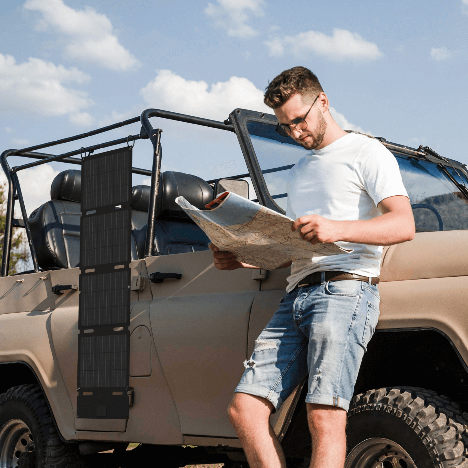 Mężczyzna w okularach przeciwsłonecznych opiera się o beżowego jeepa, patrząc na mapę. Czarny panel słoneczny jest zamontowany na drzwiach. Błękitne niebo w tle.