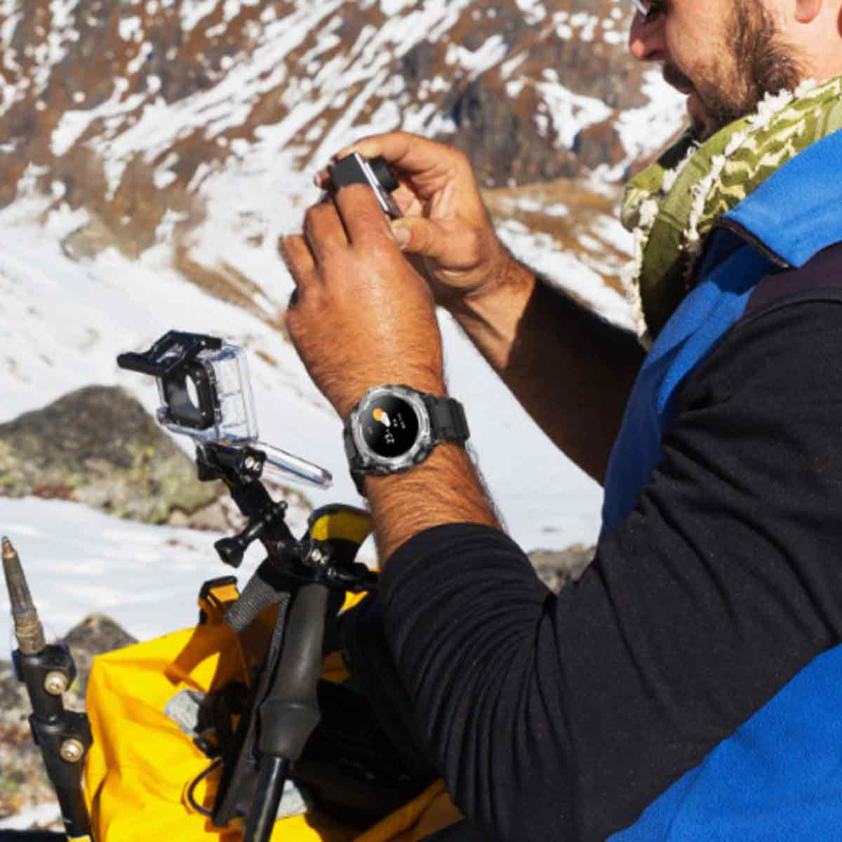 Mann mit Uhr, bedient Kamera in der Nähe eines schneebedeckten Berges. Gelbe Tasche, Kamerahalterung und Trekkingstock sichtbar.