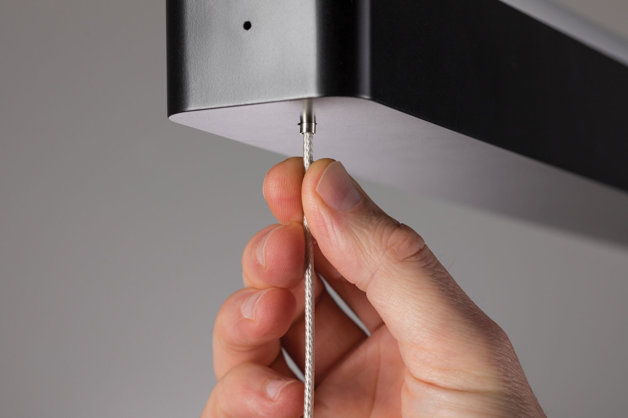 Eine Hand hält ein silbernes Kabel, das an einer schwarzen rechteckigen Vorrichtung befestigt ist. Der Hintergrund ist hellgrau.