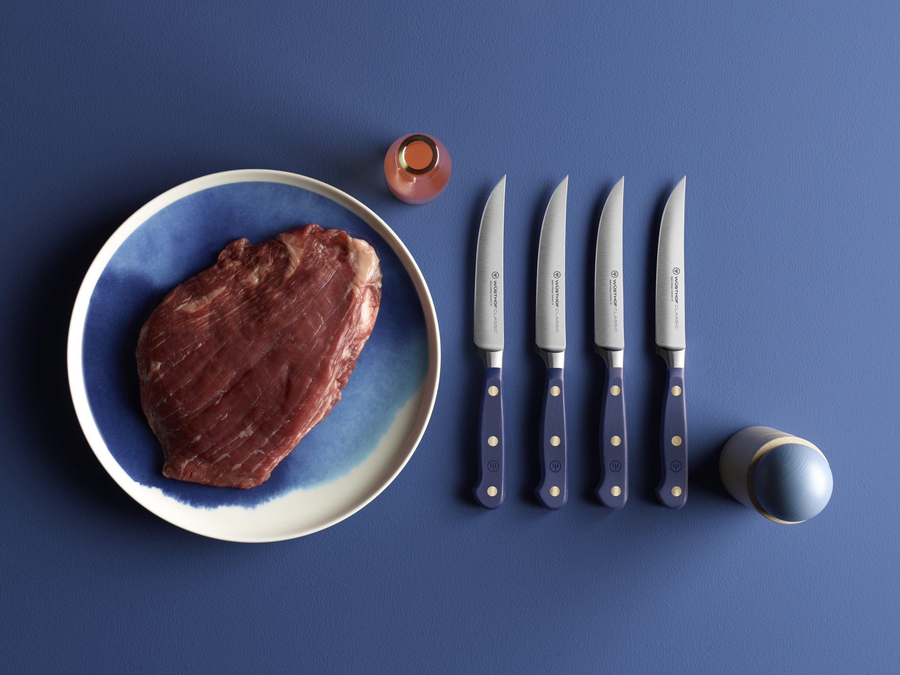 Ein rohes Steak auf einem blauen Teller, vier Messer, eine kleine Flasche und ein blaues und Holzobjekt auf einer blauen Oberfläche.