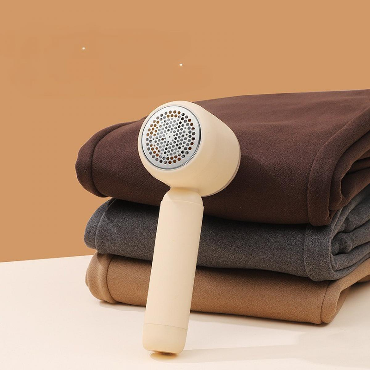 Ein Stoffrasierer liegt auf gefalteten braunen, grauen und hellbraunen Stoffen. Der Rasierer ist beige. Der Hintergrund ist orange-braun.