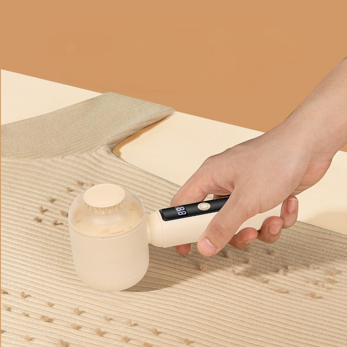 Eine Hand hält einen Fusselentferner. Das Gerät ist beige mit digitaler Anzeige. Es liegt auf einem beigen Teppich. Der Hintergrund ist hellbraun.