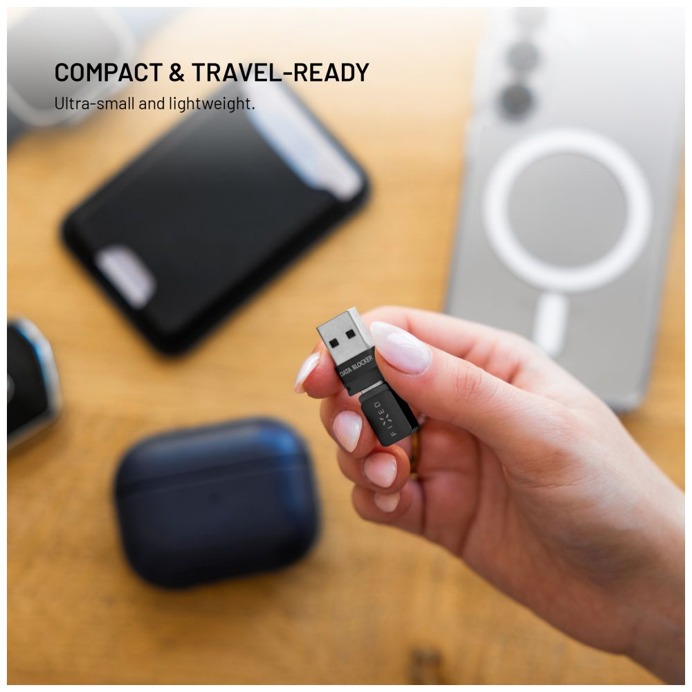 Eine Hand hält einen schwarzen USB-Datenblocker. Weitere Gegenstände sind ein Telefon, eine kabellose Ohrhörerhülle und ein Telefon mit einem MagSafe-Ladegerät.