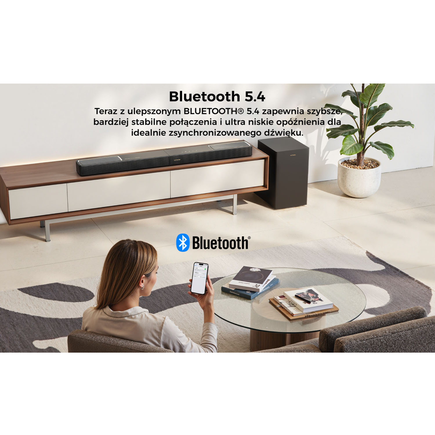 Kobieta używa telefonu do sterowania soundbarem. Soundbar i subwoofer na szafce RTV, roślina w tle. Nowoczesne wnętrze.