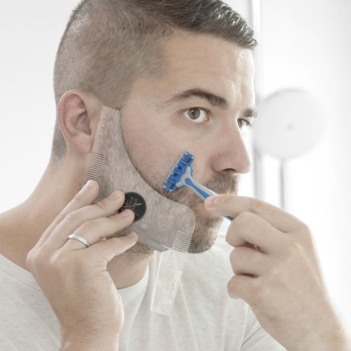 Ein Mann rasiert seinen Bart mit einem blauen Rasierer. Er benutzt ein durchsichtiges Bartformwerkzeug. Er trägt ein weißes Hemd und hat kurzes Haar.