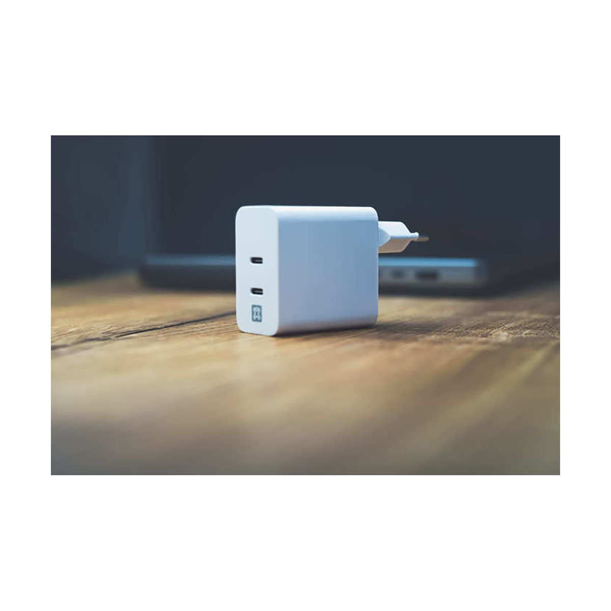Un caricabatterie bianco è su un tavolo di legno. Un laptop è sullo sfondo. Il caricabatterie ha due porte USB-C e una spina.